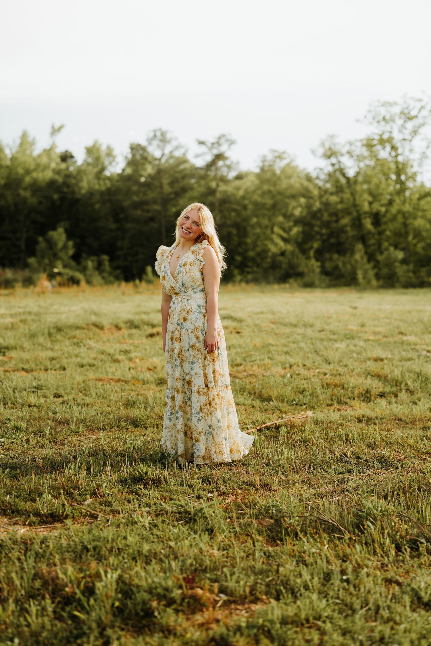 A blonde woman in a floral maxi dress stands smiling in a green field with trees in the background.