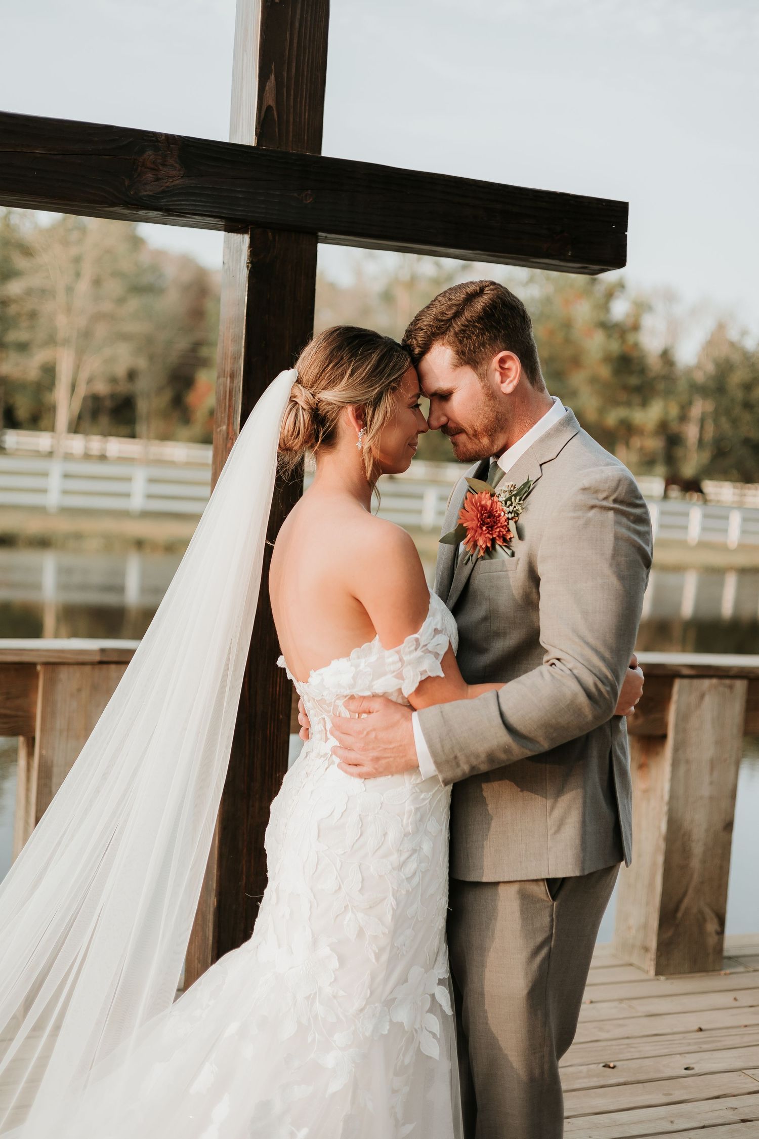 A romantic wedding photo of a couple embracing under a wooden cross on a rustic deck.