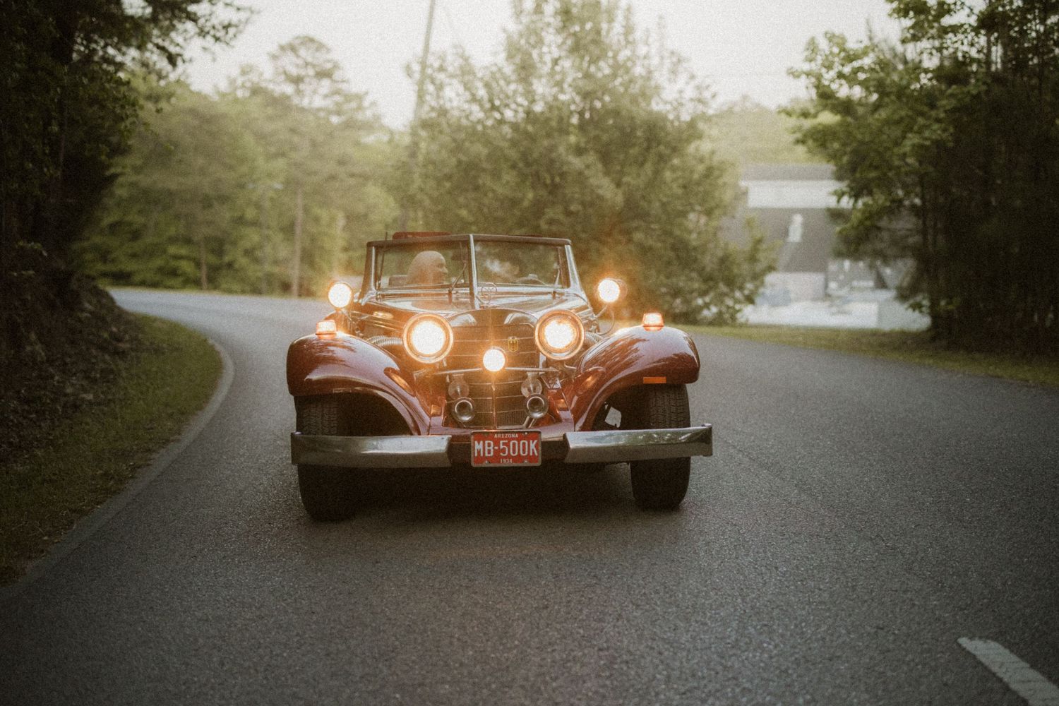 Vintage classic car with headlights on driving down a tree-lined road at dusk.