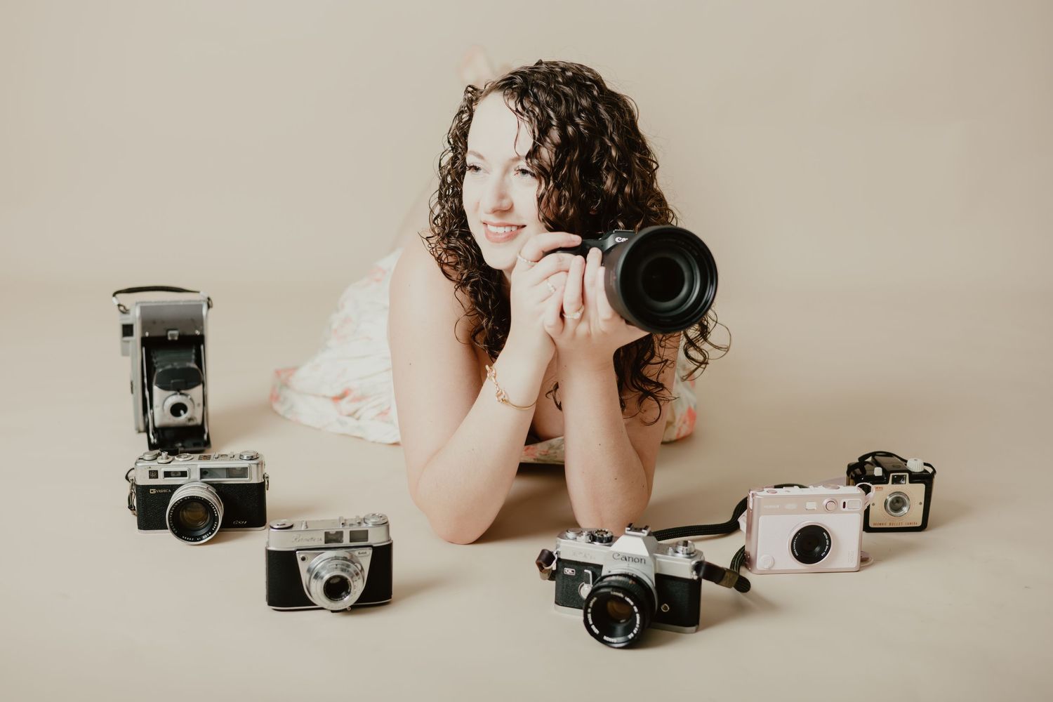 Young woman with curly hair poses with a DSLR camera surrounded by vintage cameras on a beige studio floor.