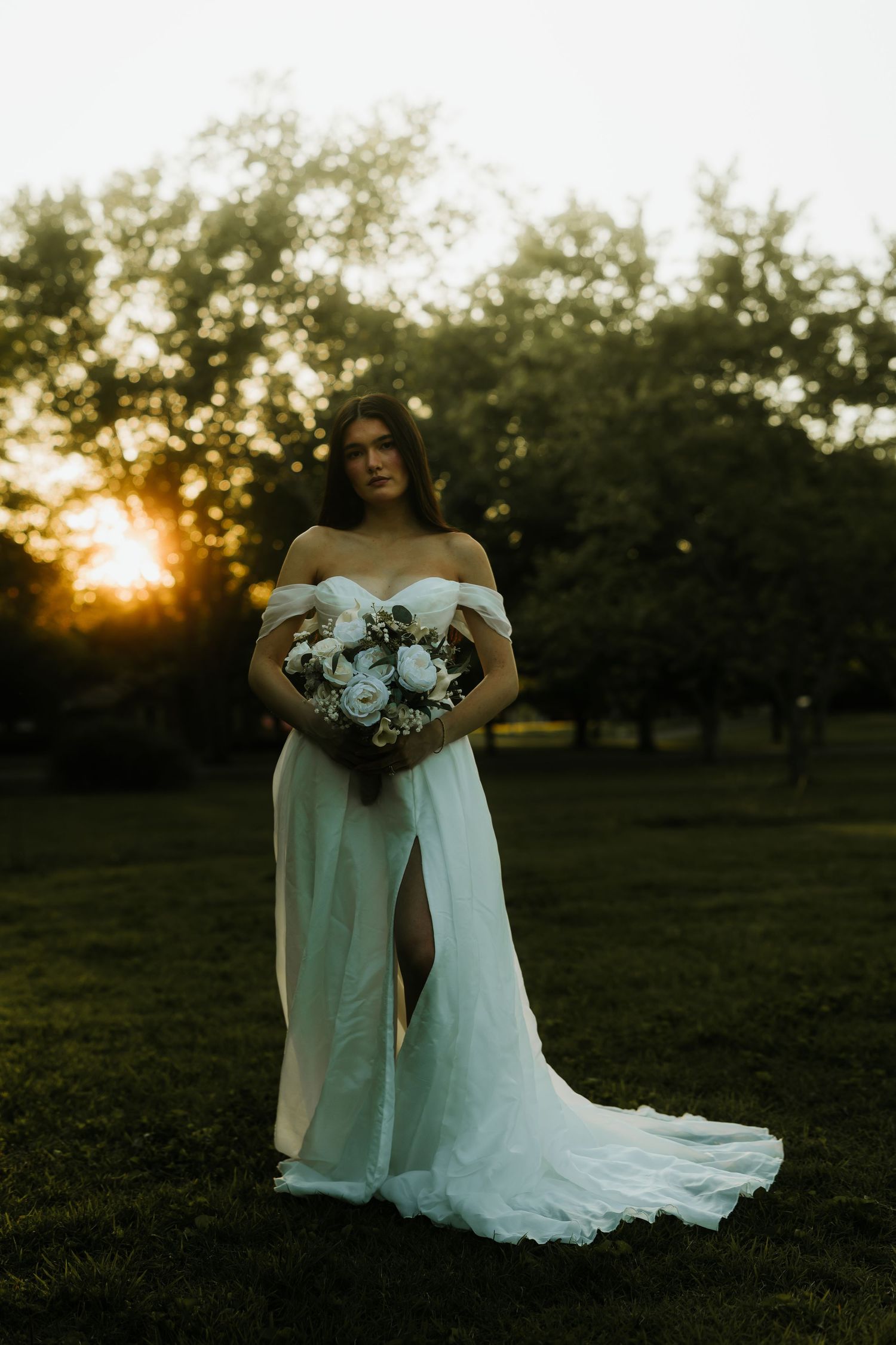 Bride in off-shoulder white gown with slit holds bouquet at golden sunset in lush green garden.