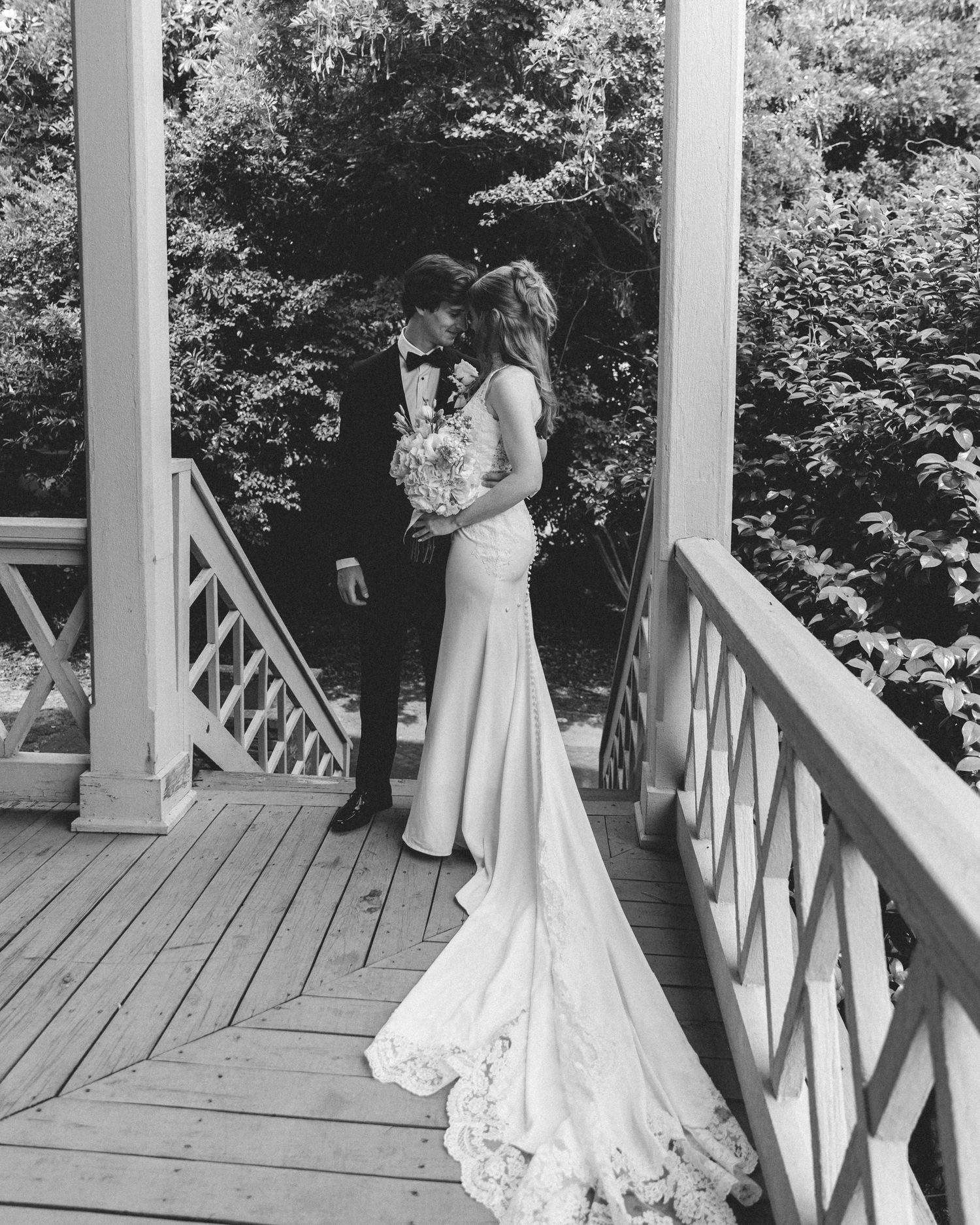 Black and white wedding photo of bride and groom sharing a romantic moment on a wooden gazebo bridge surrounded by trees.