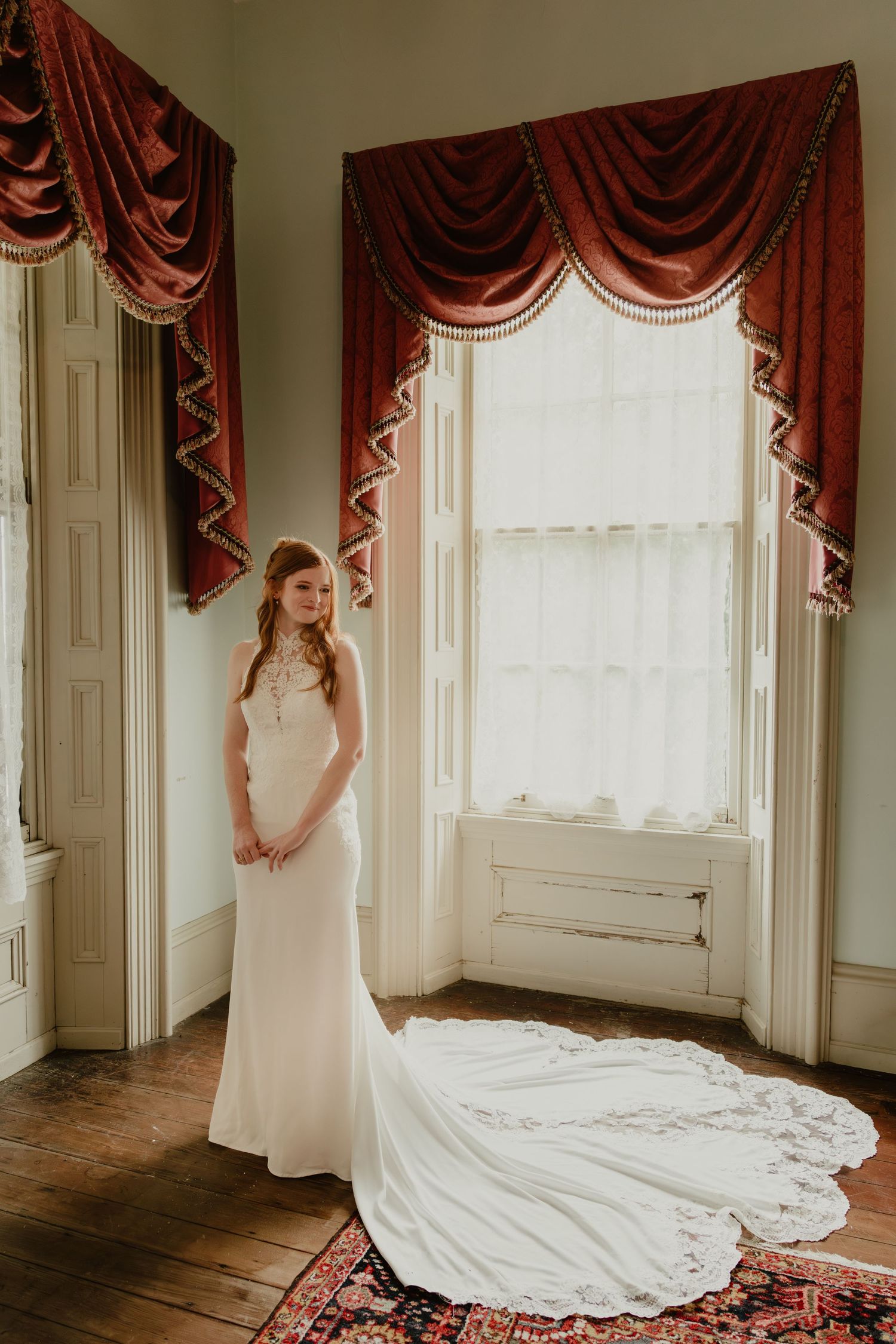 Bride in elegant white gown with lace train stands by bay window with red velvet drapes on hardwood floor.