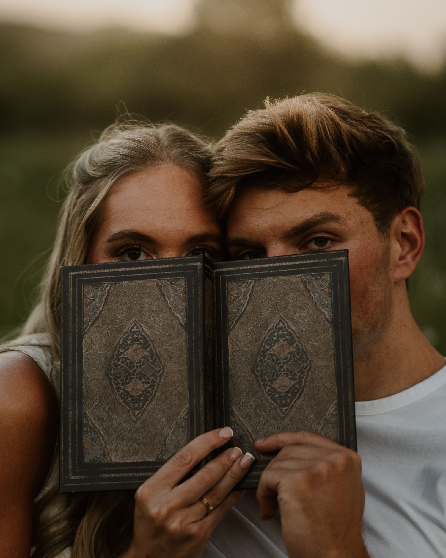Couple holding an ornate vintage book covering their faces during a romantic outdoor engagement photo session.