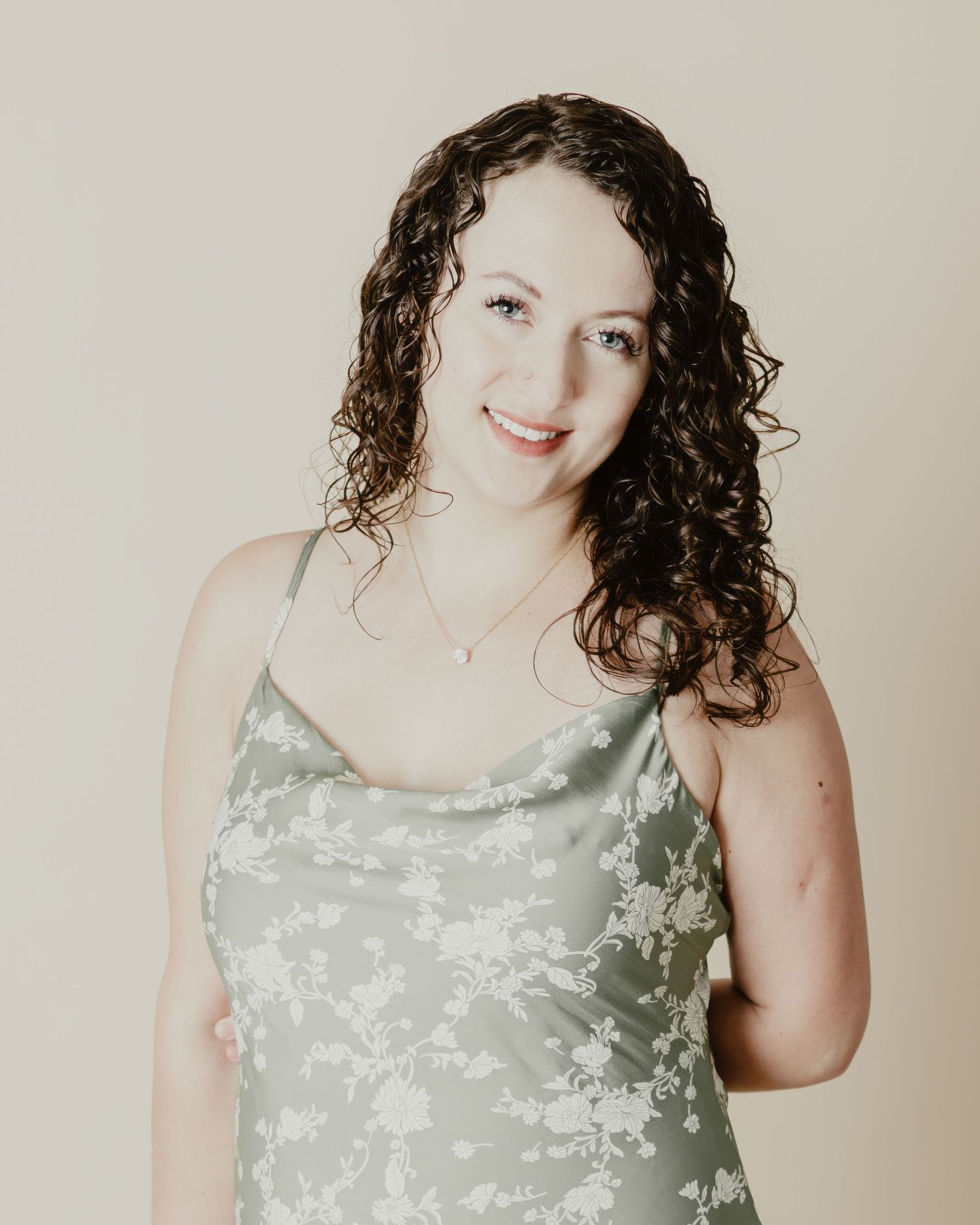 Woman with curly dark hair smiling, wearing a sage green floral satin slip dress against a neutral background.