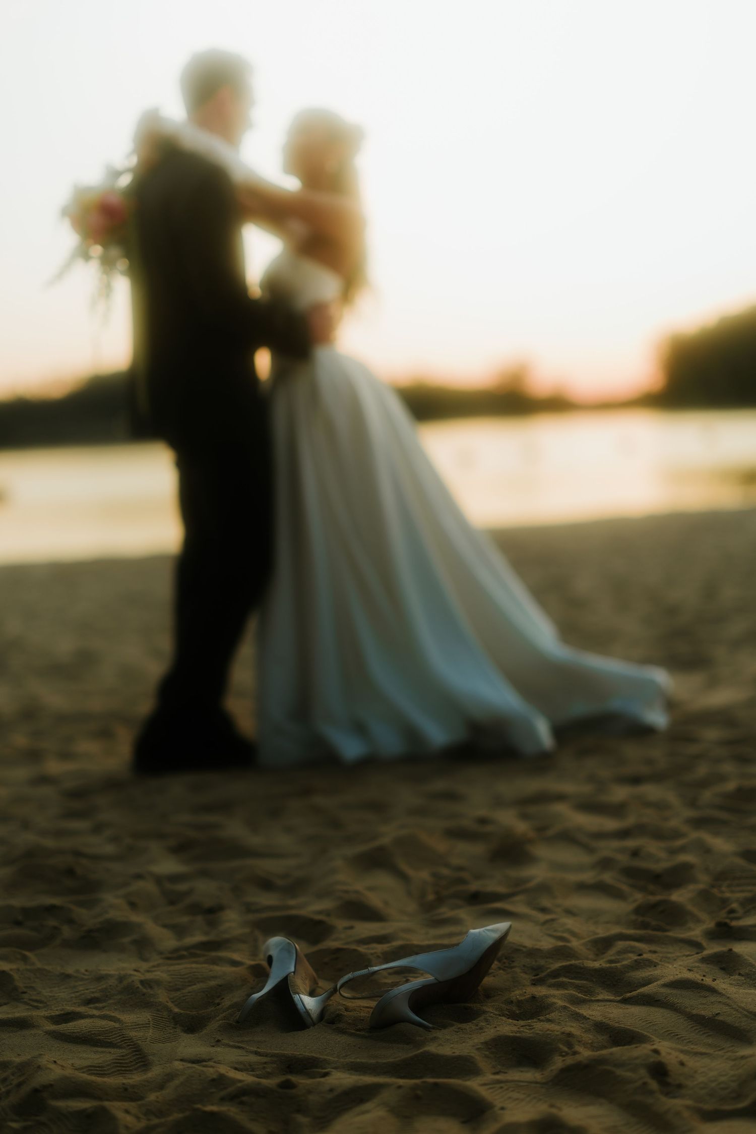 Bride and groom embrace on sandy beach at sunset with silver heels in foreground.
