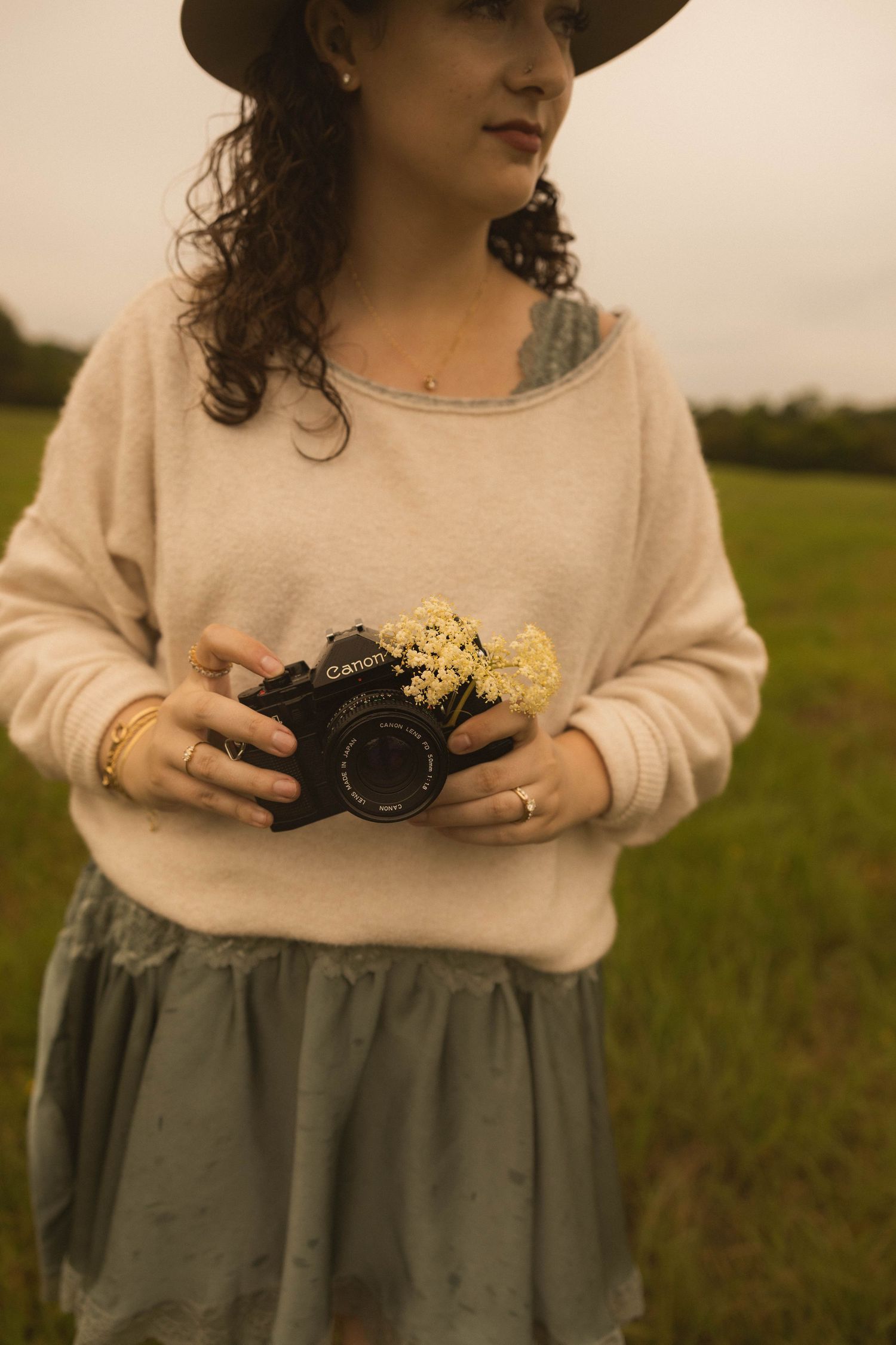 Woman in hat and cream sweater holding a Canon camera with small yellow wildflowers in a green field.