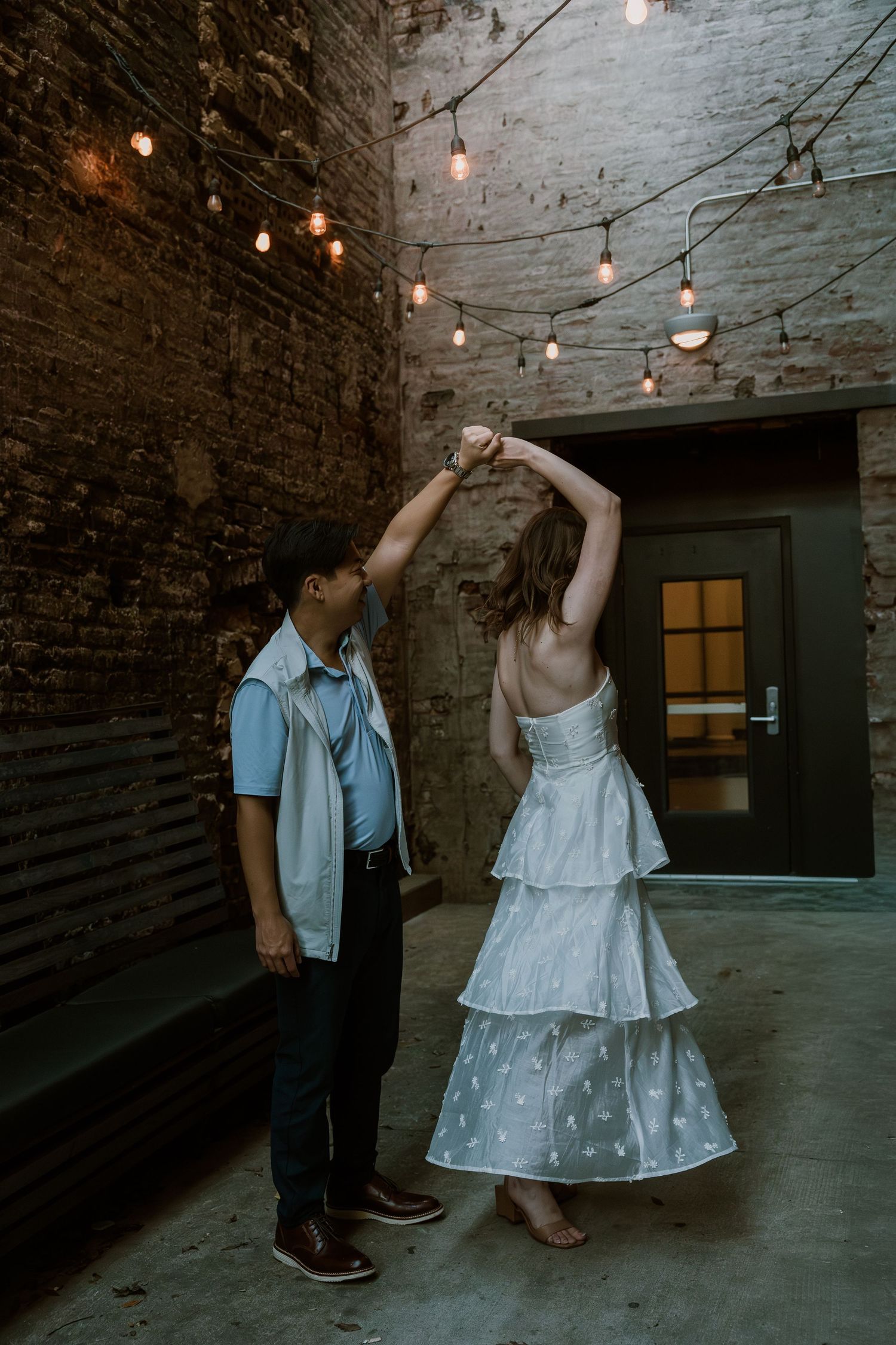 A couple dancing together in a rustic brick-walled venue decorated with string lights.