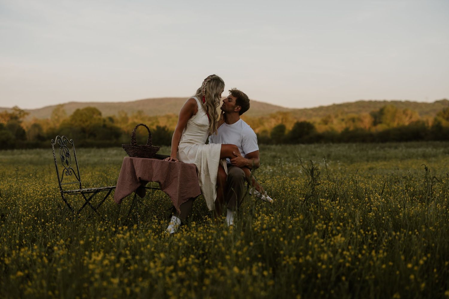 Couple sharing a romantic kiss in a golden field of wildflowers at sunset with rolling hills in the background.