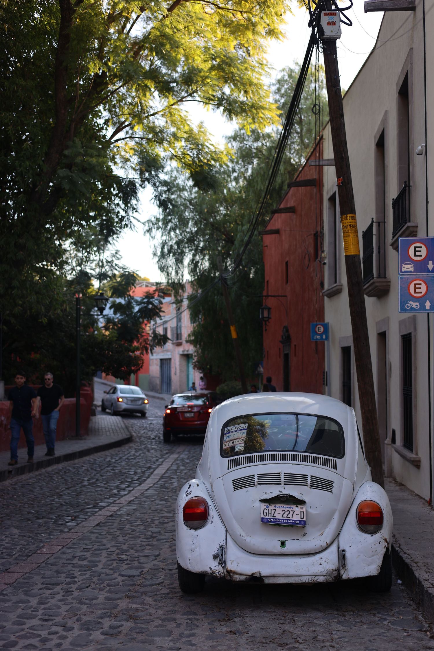 White VW Beetle parked on a cobblestone street in a colorful colonial town with trees and buildings.