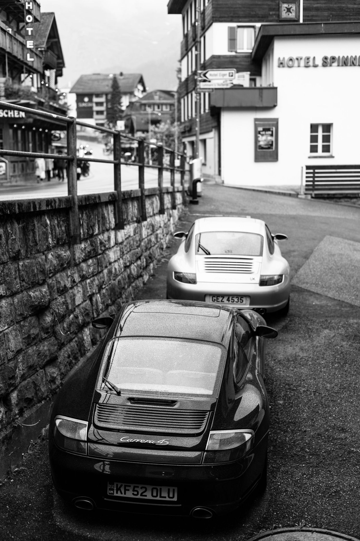 Two Porsche sports cars parked on a narrow alpine village street in black and white.