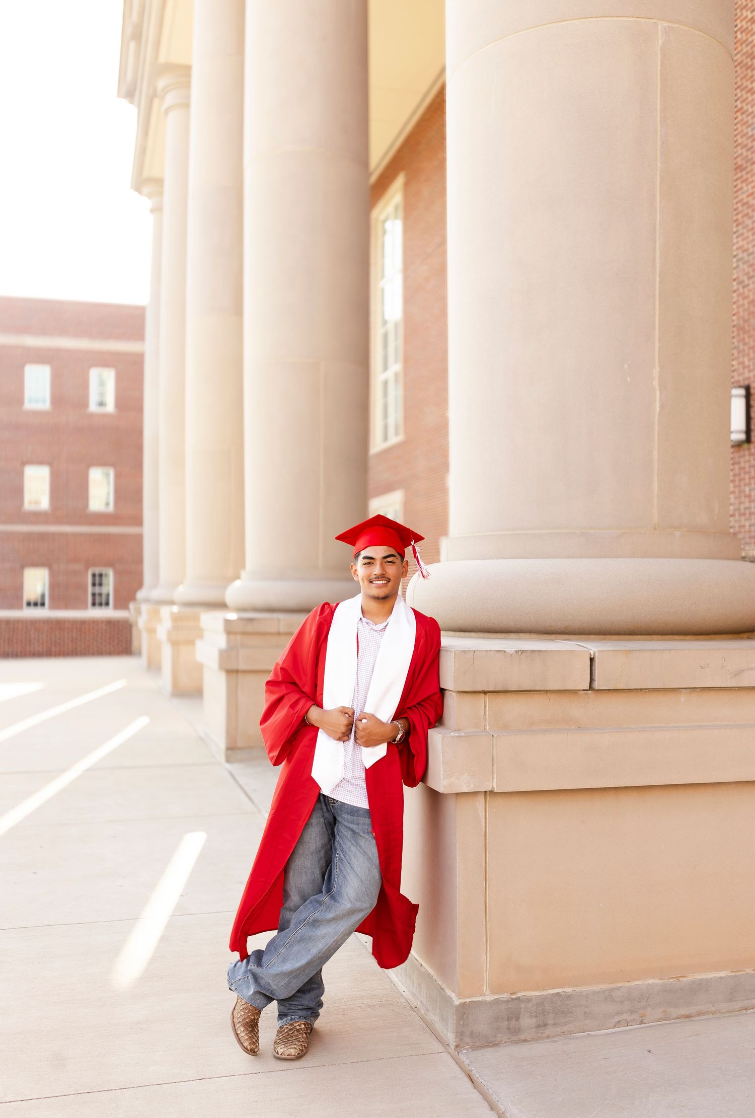 Smiling male graduate in red cap and gown leaning against large columns outdoors.