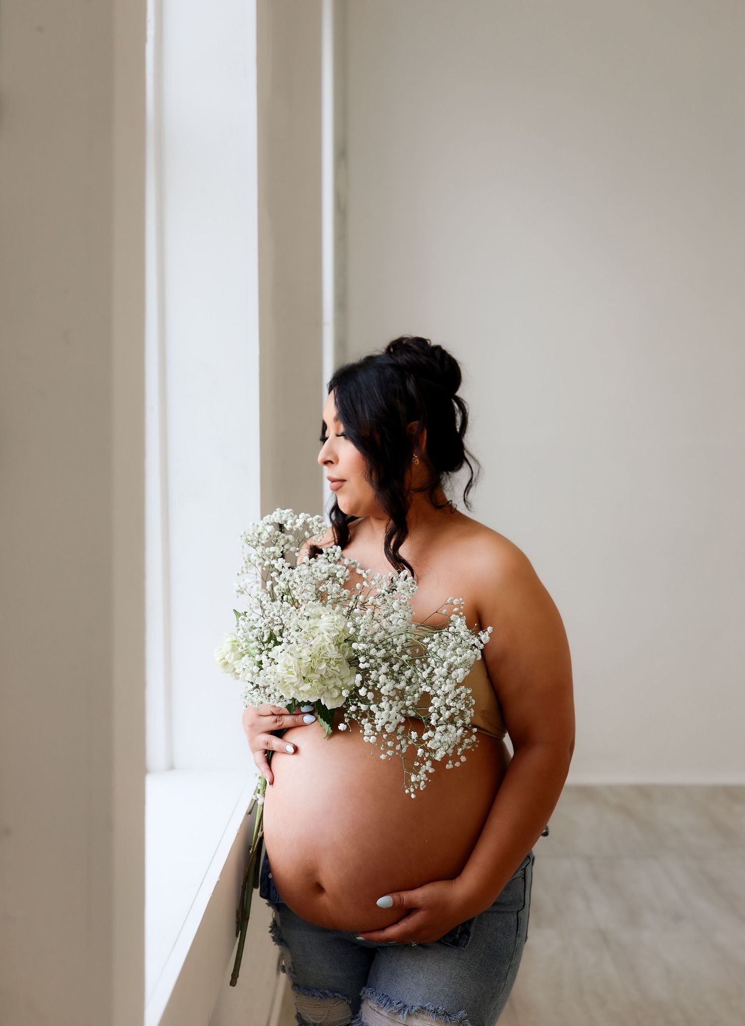 Pregnant woman holding white baby's breath bouquet by window in soft natural light maternity photo.