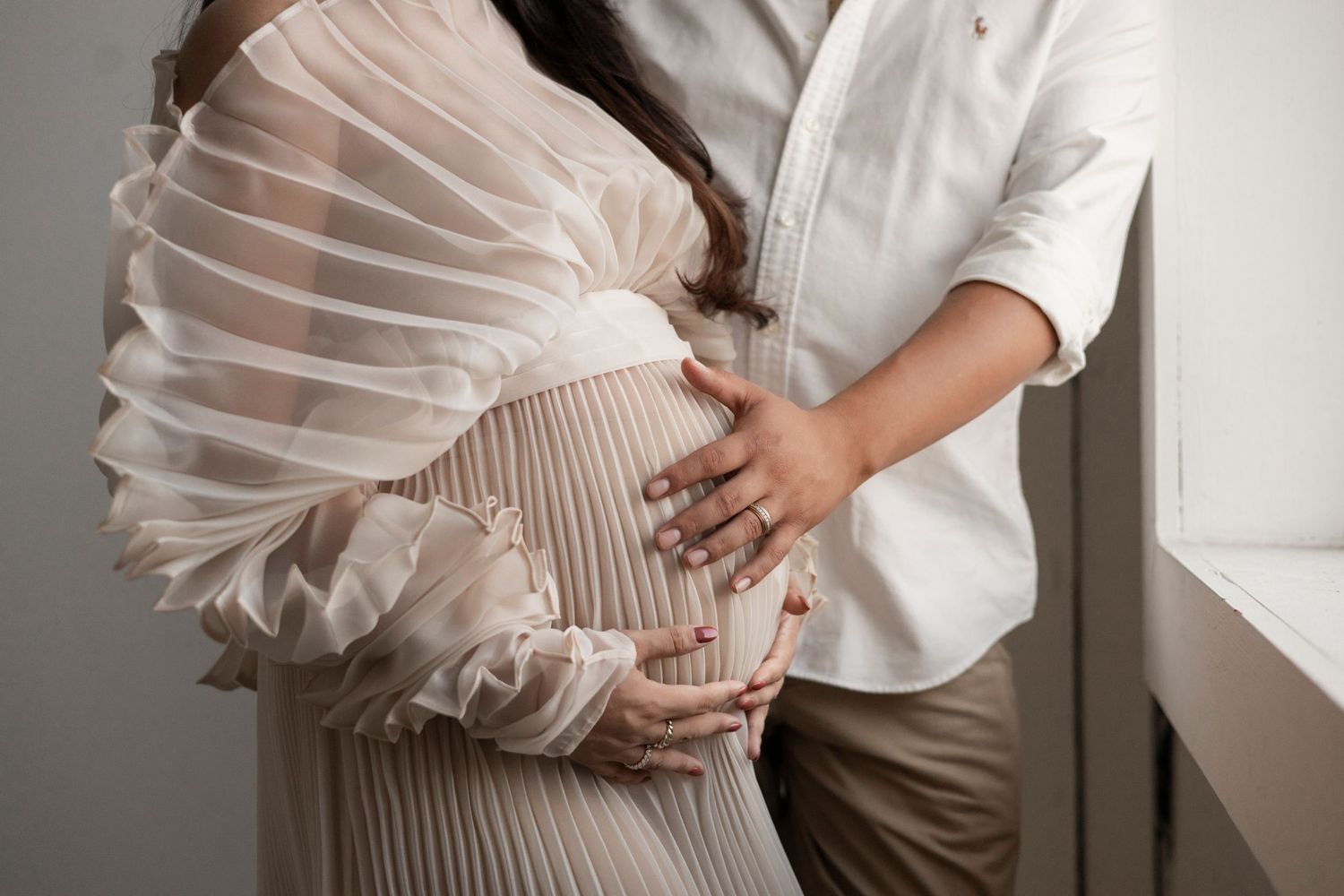 Close-up of couple tenderly cradling pregnant belly, woman in sheer pleated dress, partner in white shirt.
