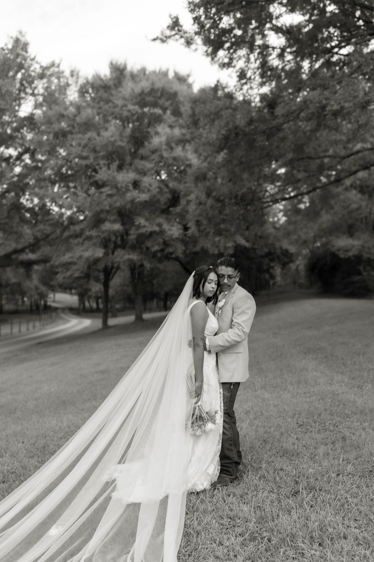 Black and white photo of bride and groom embracing outdoors with flowing cathedral veil trailing across green lawn.