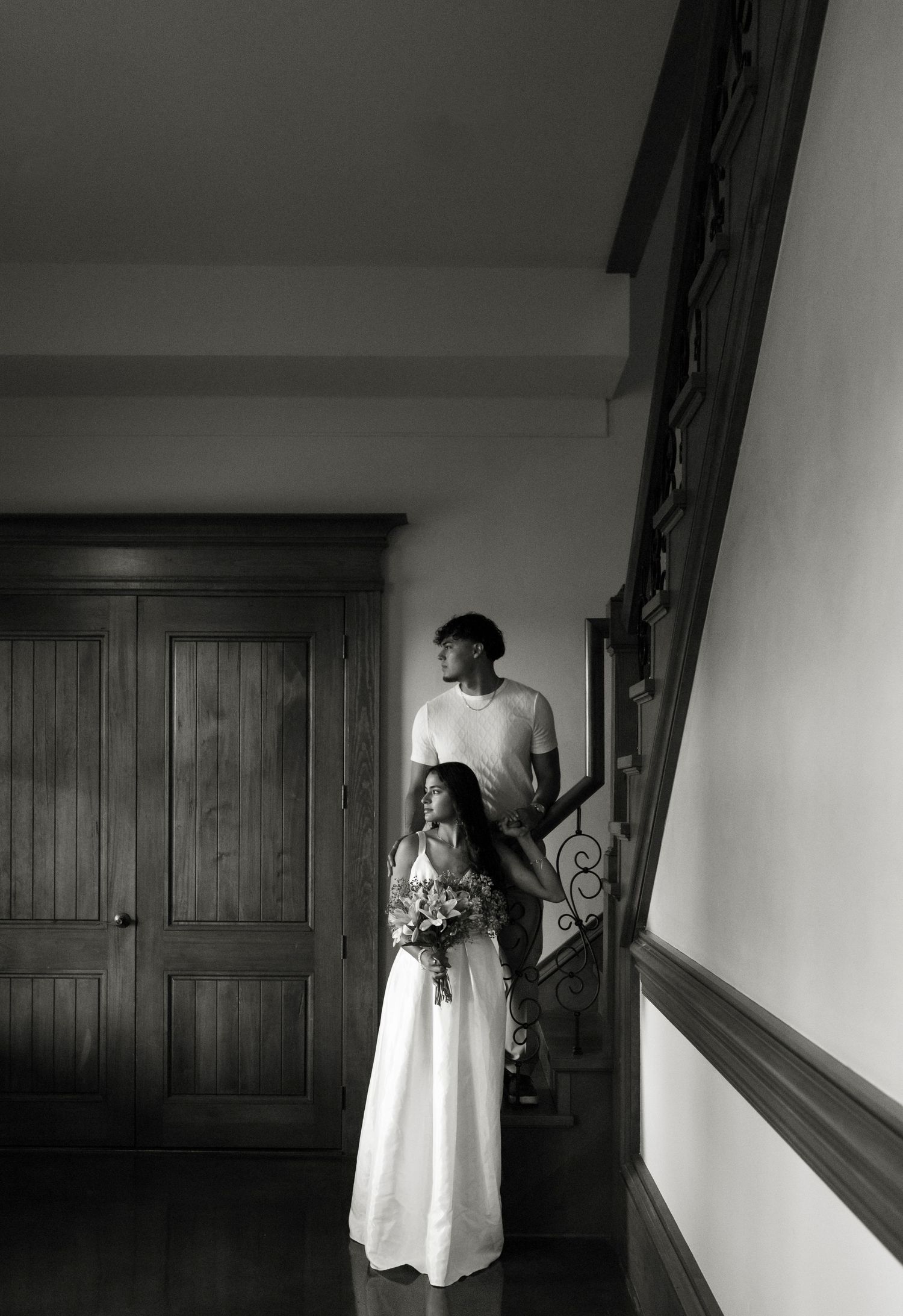 Black and white photo of couple standing by a staircase with bouquet.