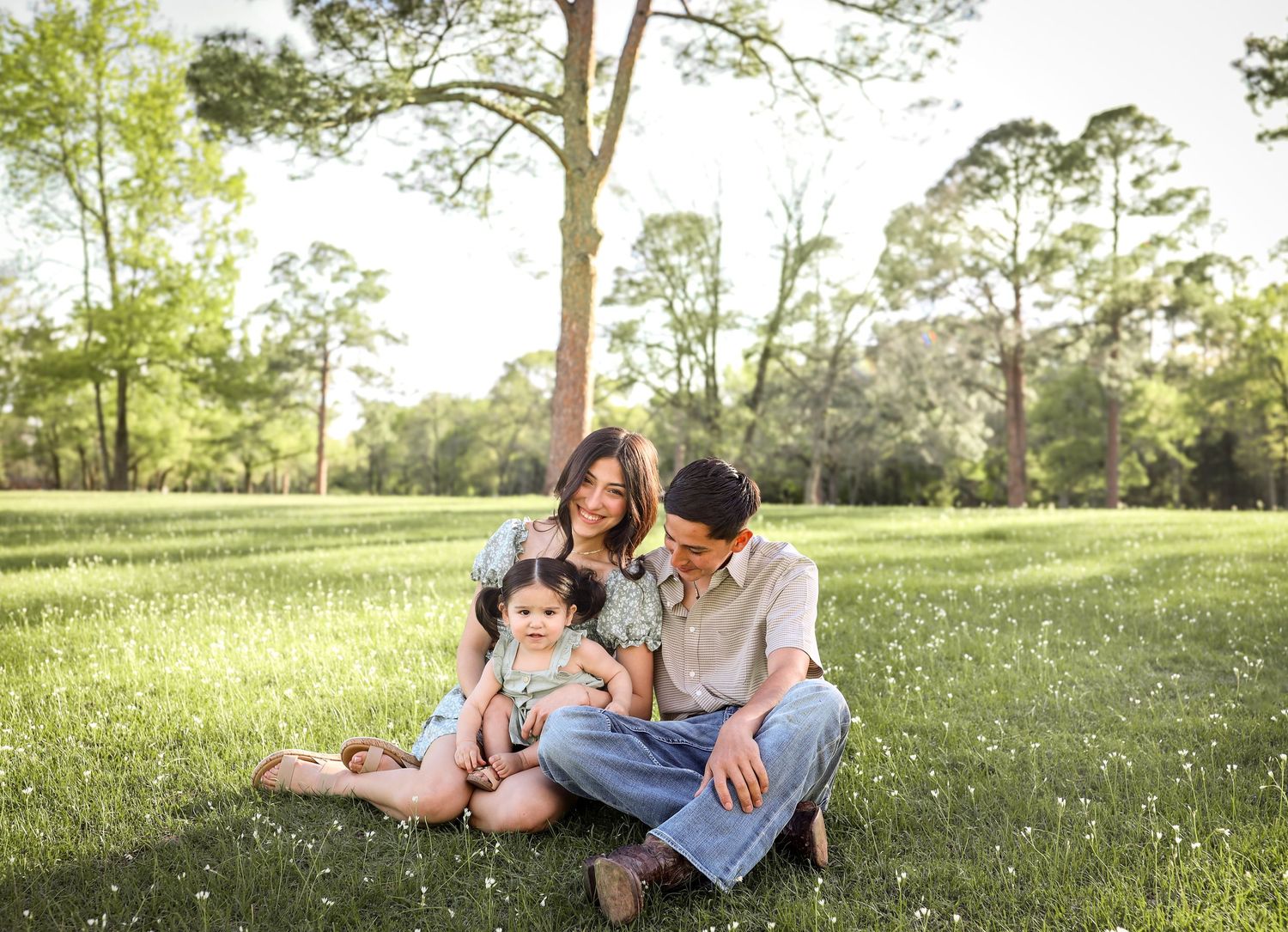 Family of four sitting together on green grass in sunlit park, parents smiling at two young children.