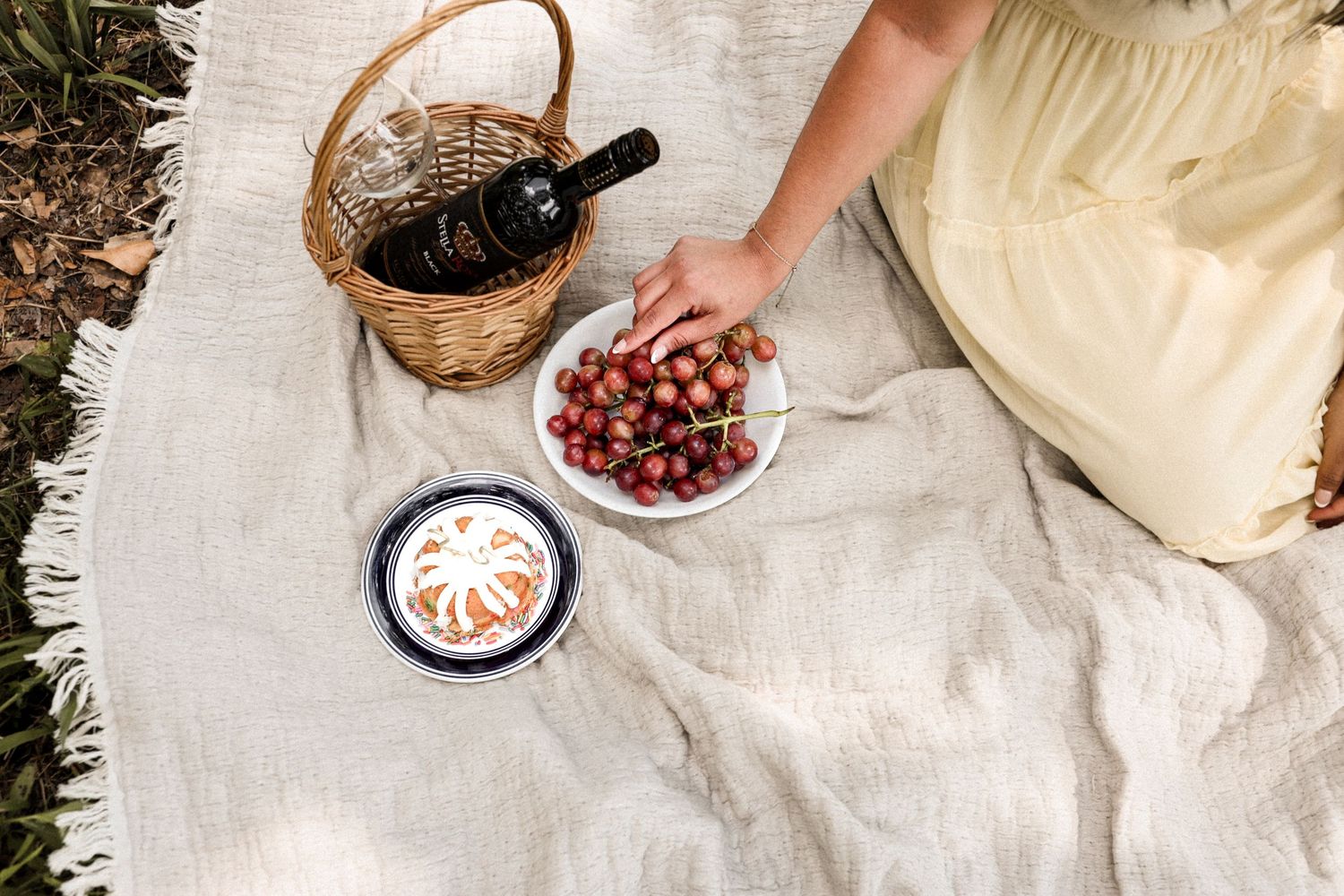 Overhead view of picnic blanket with cherries bowl, wine bottle in basket, and decorative plate outdoors.