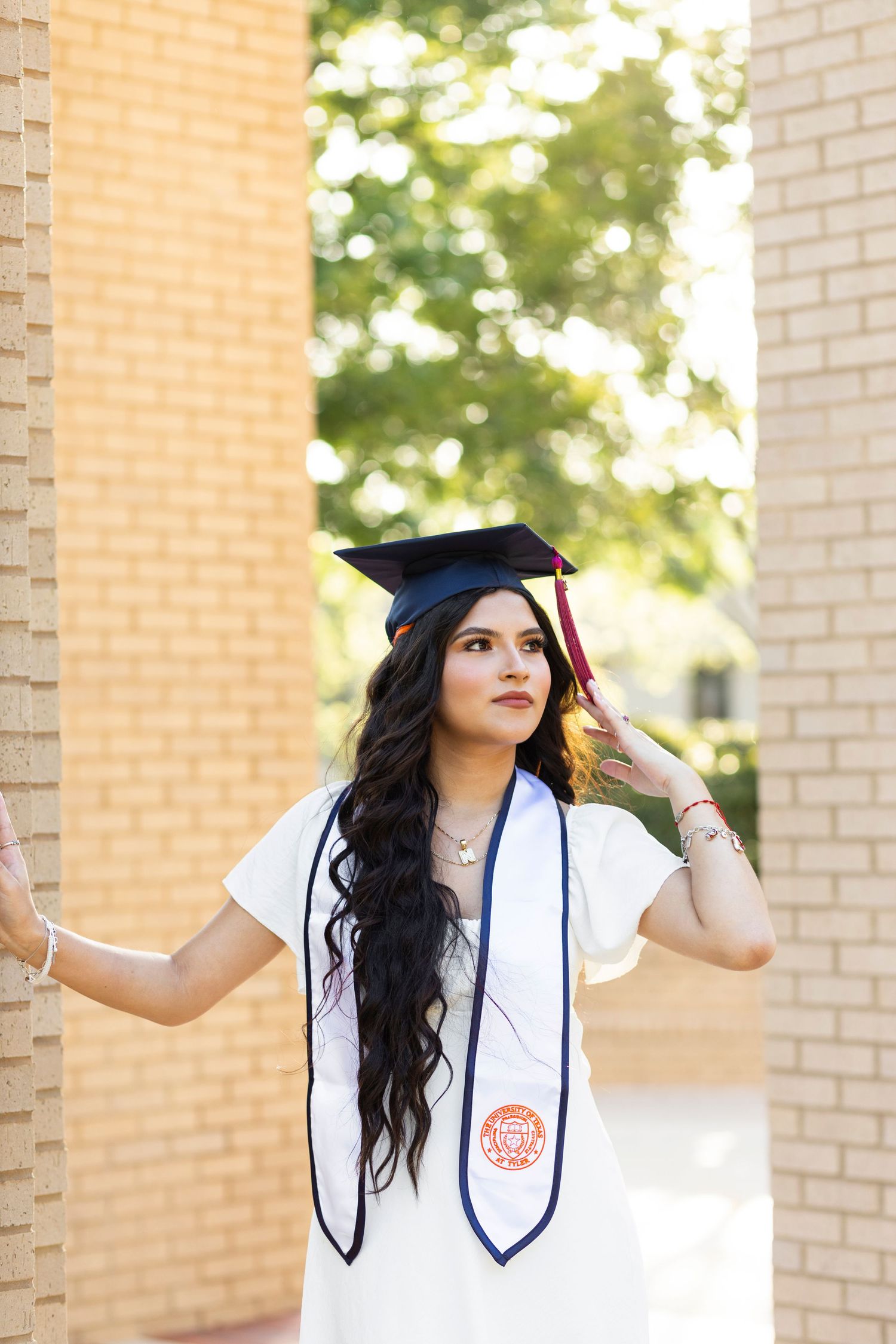 Young woman in graduation cap and white gown with stole poses between brick columns outdoors.