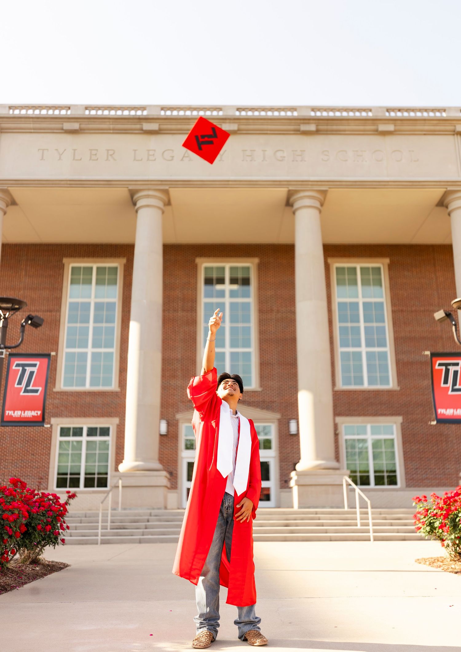 Male graduate in red gown tossing cap in air outside Tyler Legacy High School.