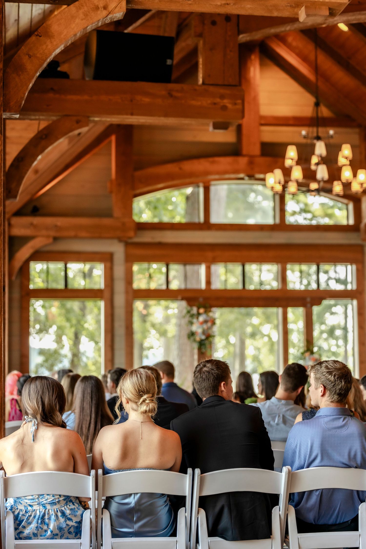 Wedding guests seated in white chairs inside a rustic timber-frame venue with warm wood beams and chandeliers.
