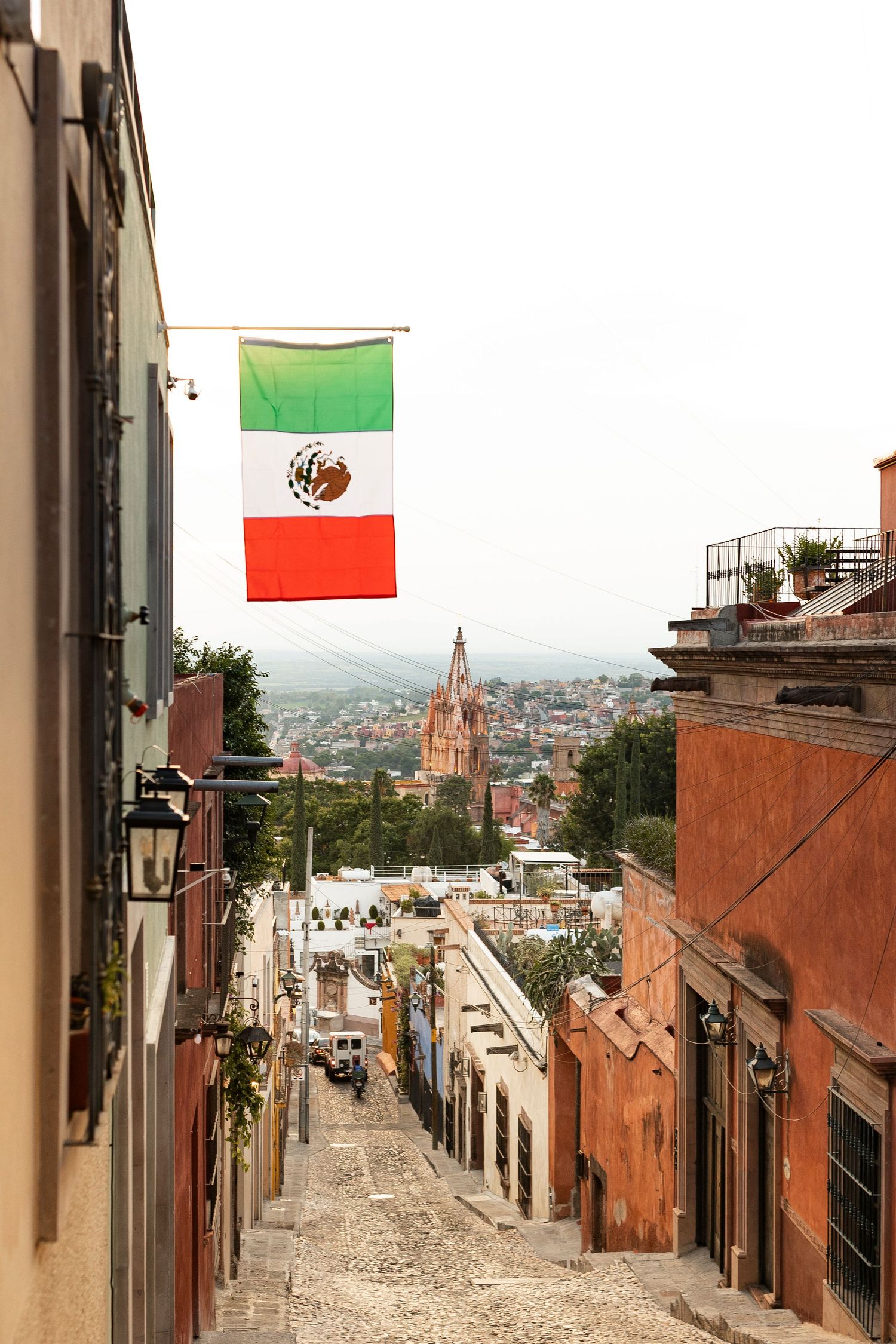 A Mexican flag hangs above a cobblestone street with a cathedral spire in background.