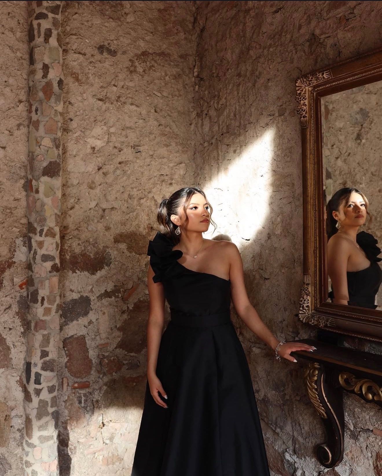 Woman in elegant black one-shoulder gown poses beside ornate mirror in rustic stone room.
