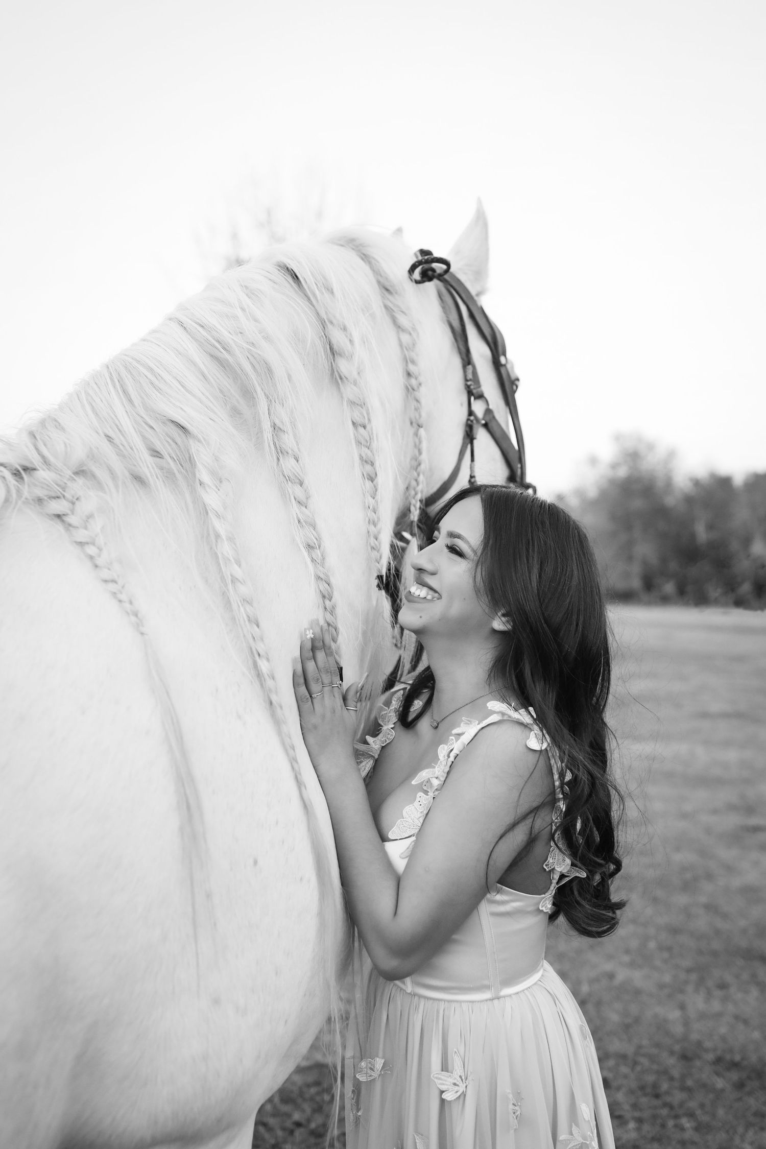 Black and white photo of smiling brunette woman in dress affectionately leaning against white braided horse.