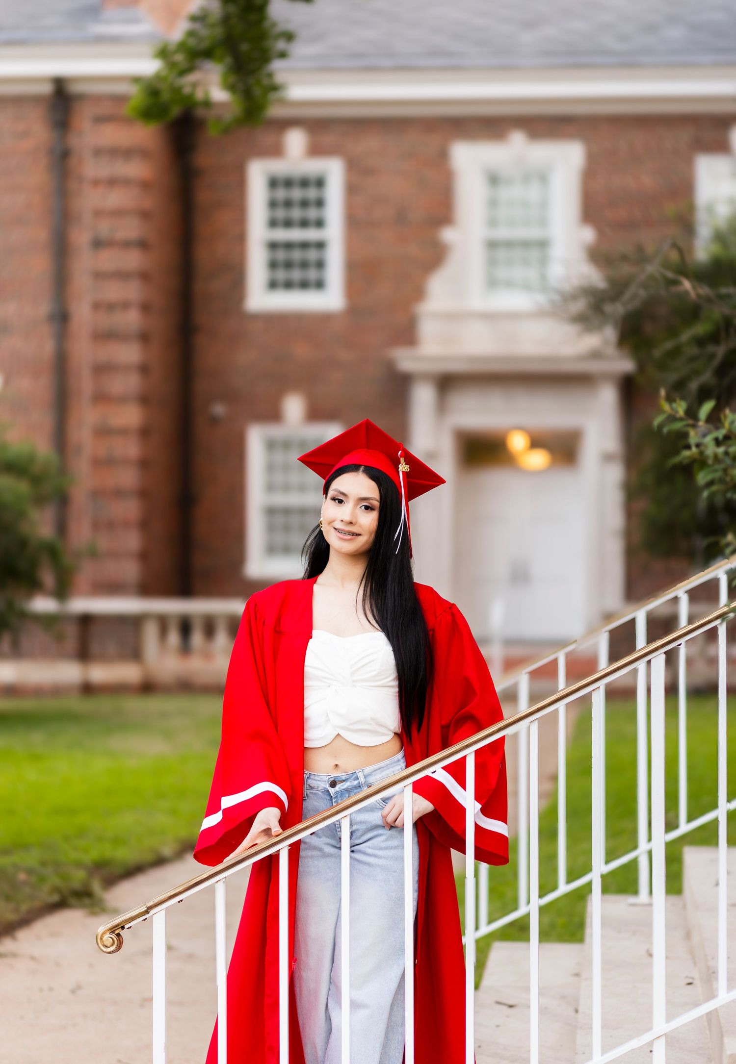 Graduate in red cap and gown poses on staircase in front of classic brick university building.