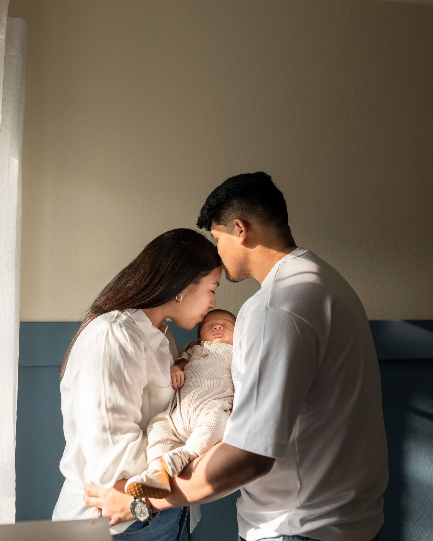 Young couple tenderly lean together over their newborn baby in warm natural light indoors.