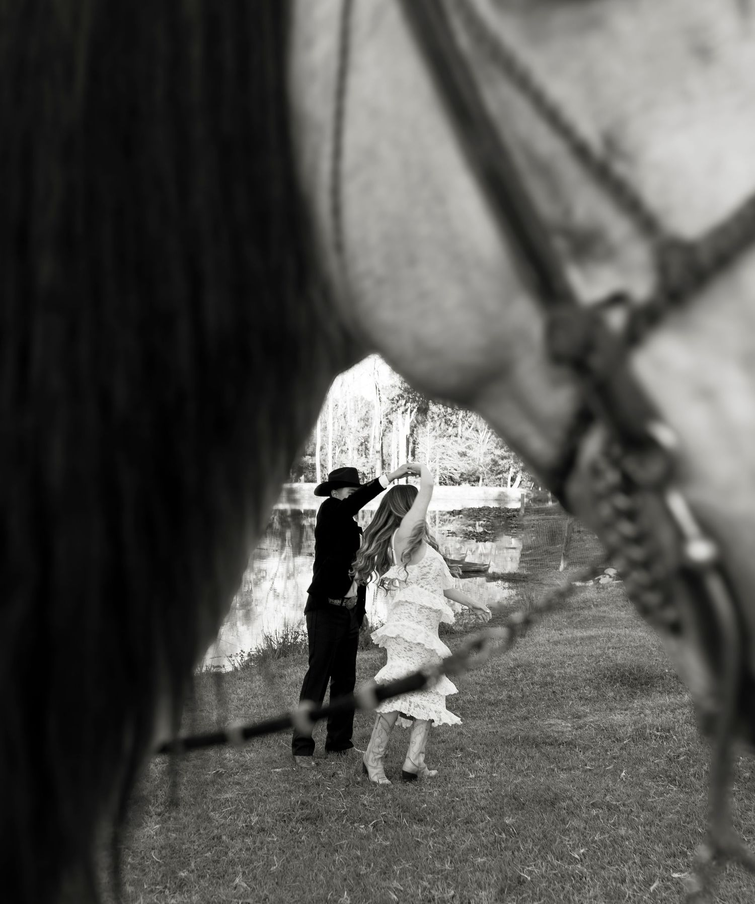 Black and white artistic photo of a couple framed between two horses near a peaceful pond setting.