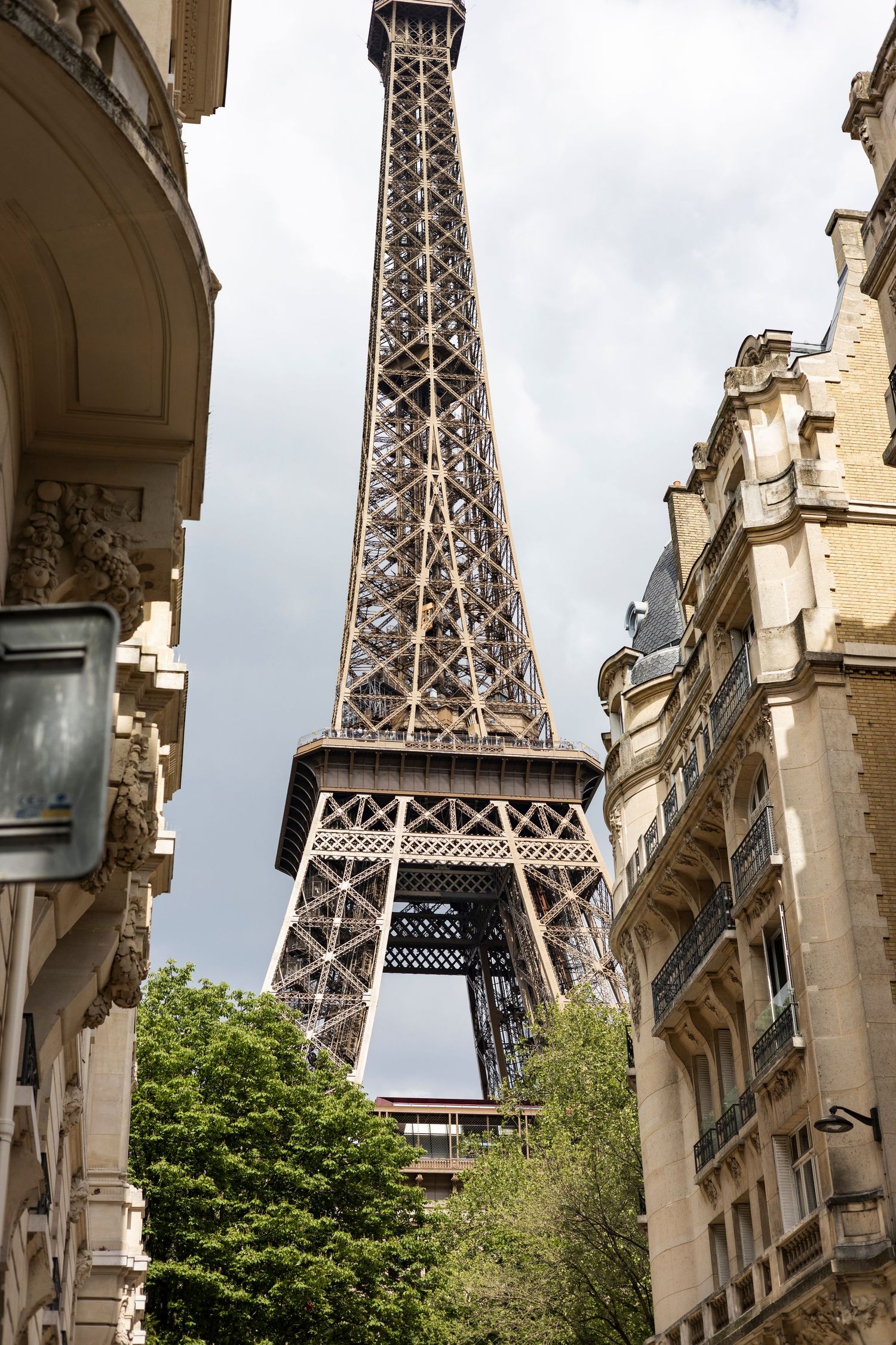 The iconic Eiffel Tower rises above Parisian buildings and green trees under cloudy skies.