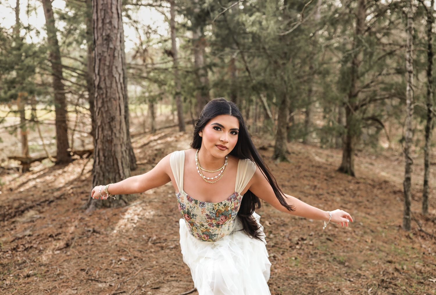 Young brunette woman in floral corset and white skirt twirling with arms outstretched in pine forest.