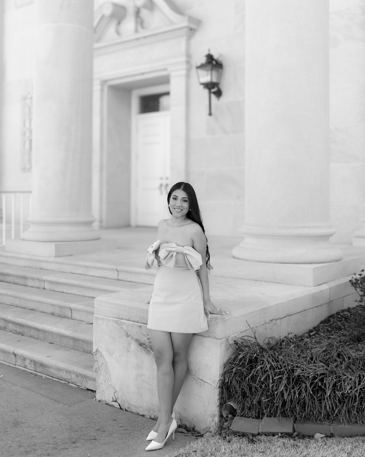 Black and white photo of smiling woman in off-shoulder mini dress posing on steps between columns.