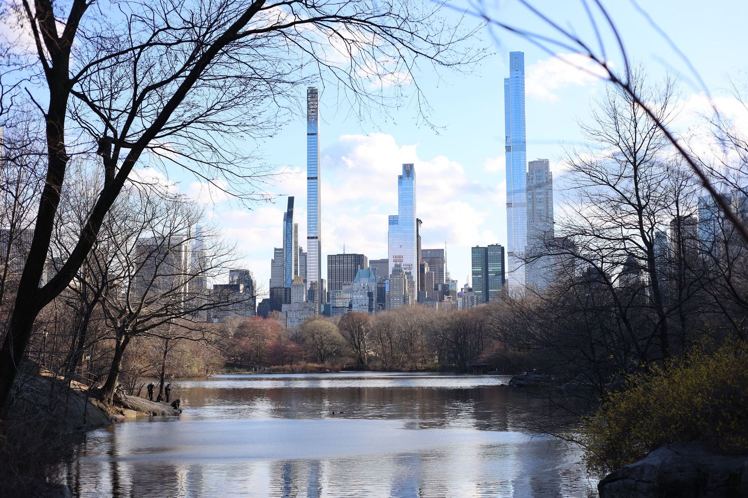 Central Park lake reflecting NYC skyline with tall skyscrapers and bare winter trees on a clear sunny day.