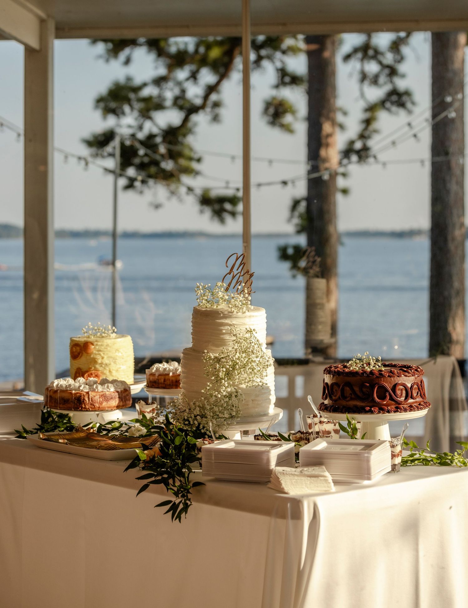 Elegant wedding cake display with multiple tiered cakes and greenery set against a scenic lakeside window.