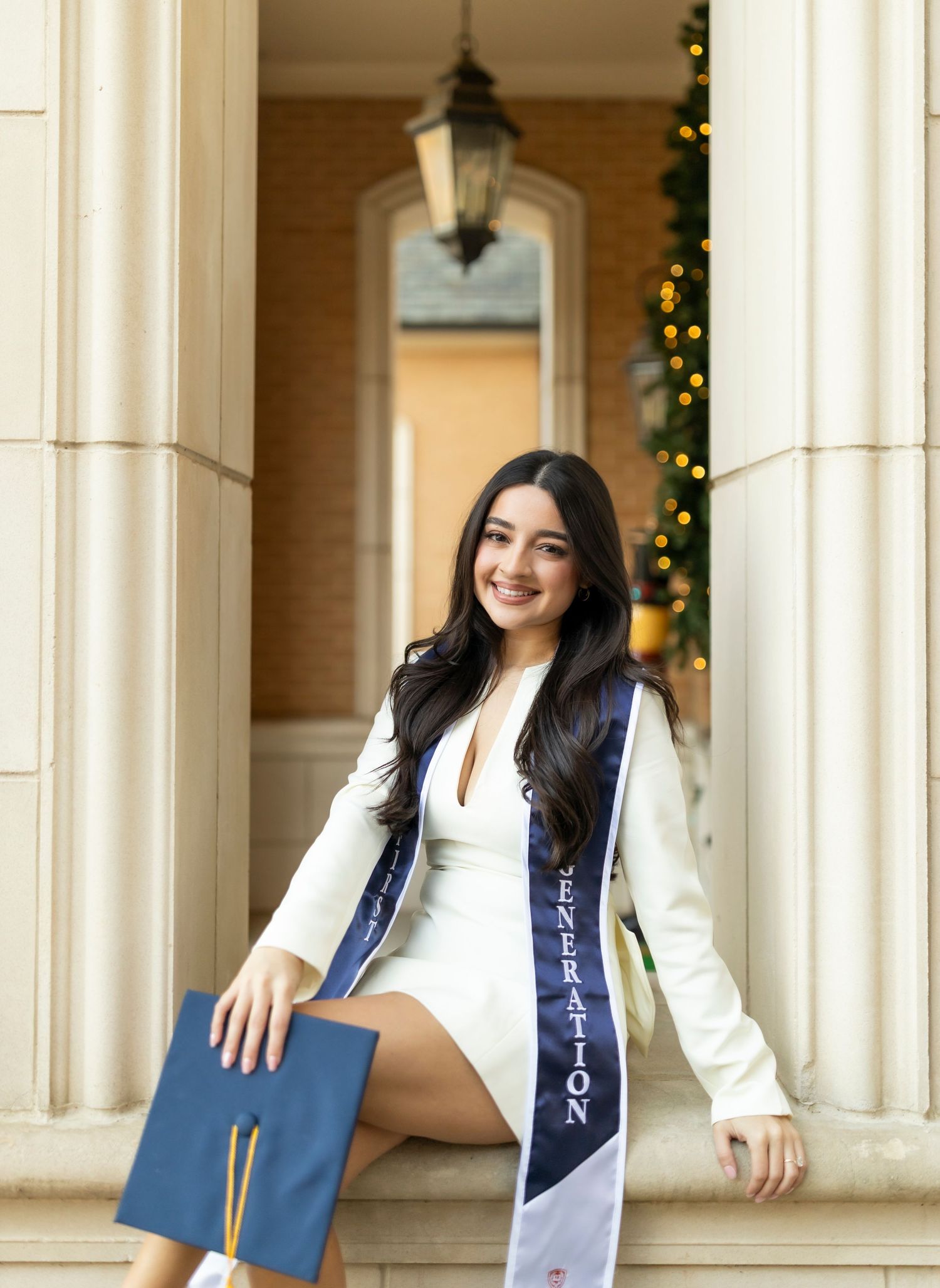 Female graduate in white dress with navy graduation sash sits on steps holding cap.