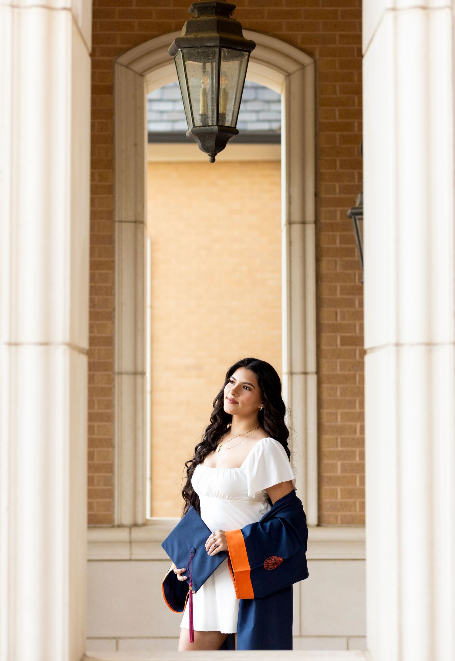 Graduate in white dress holds navy and orange regalia beside elegant column with lantern backdrop.