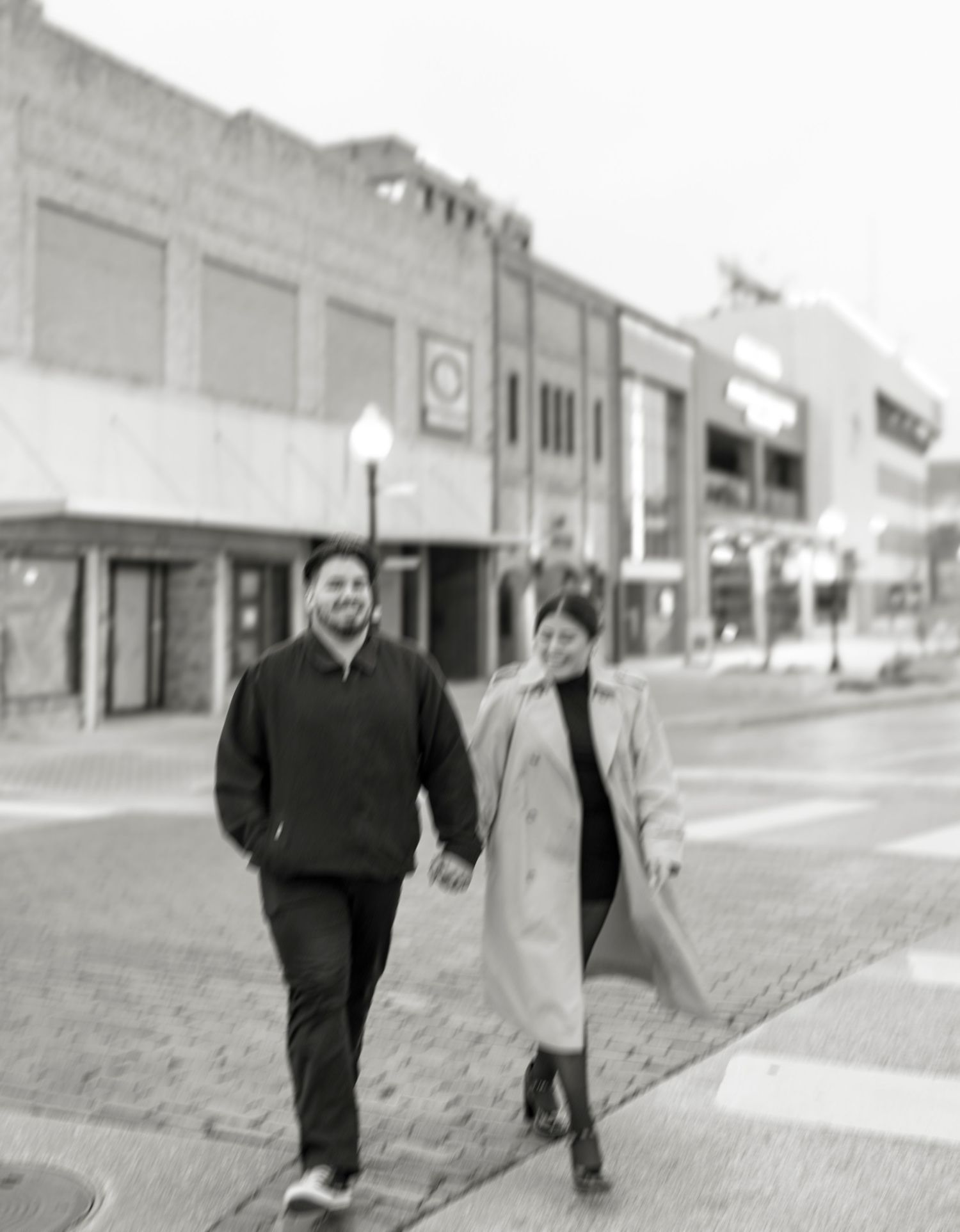 Black and white photo of a couple walking hand in hand along a downtown city sidewalk past storefronts.
