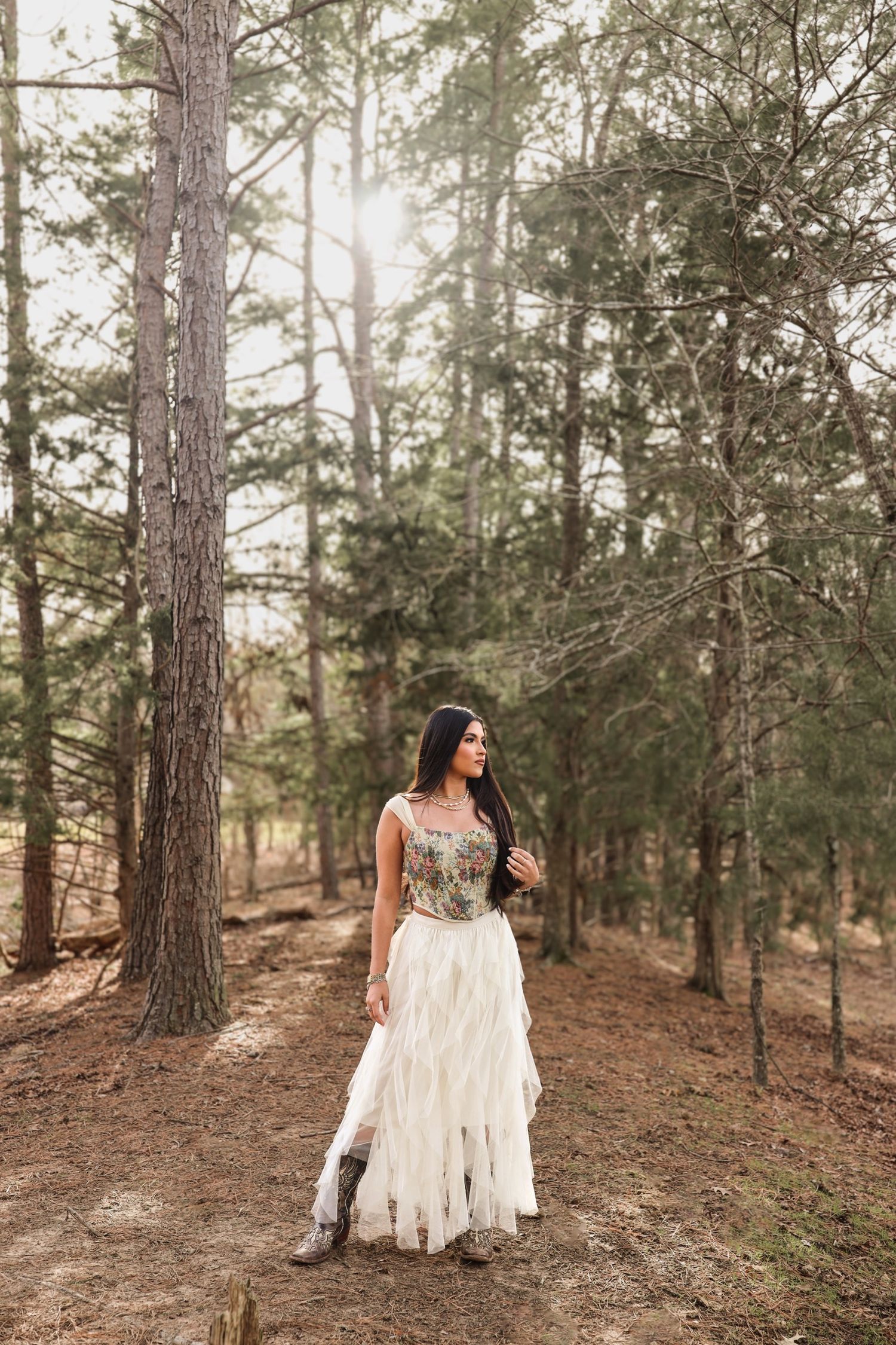 Brunette woman in white ruffled dress and boots standing among tall pine trees in sunlit forest.