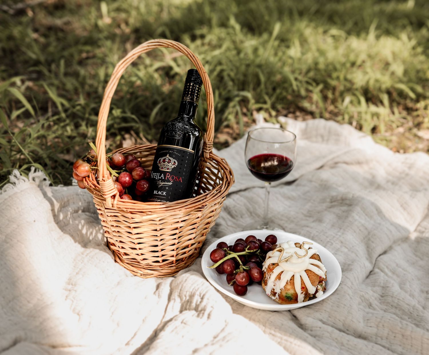 Wicker picnic basket with wine bottle, fresh strawberries, cheese plate, and red wine glass on blanket.