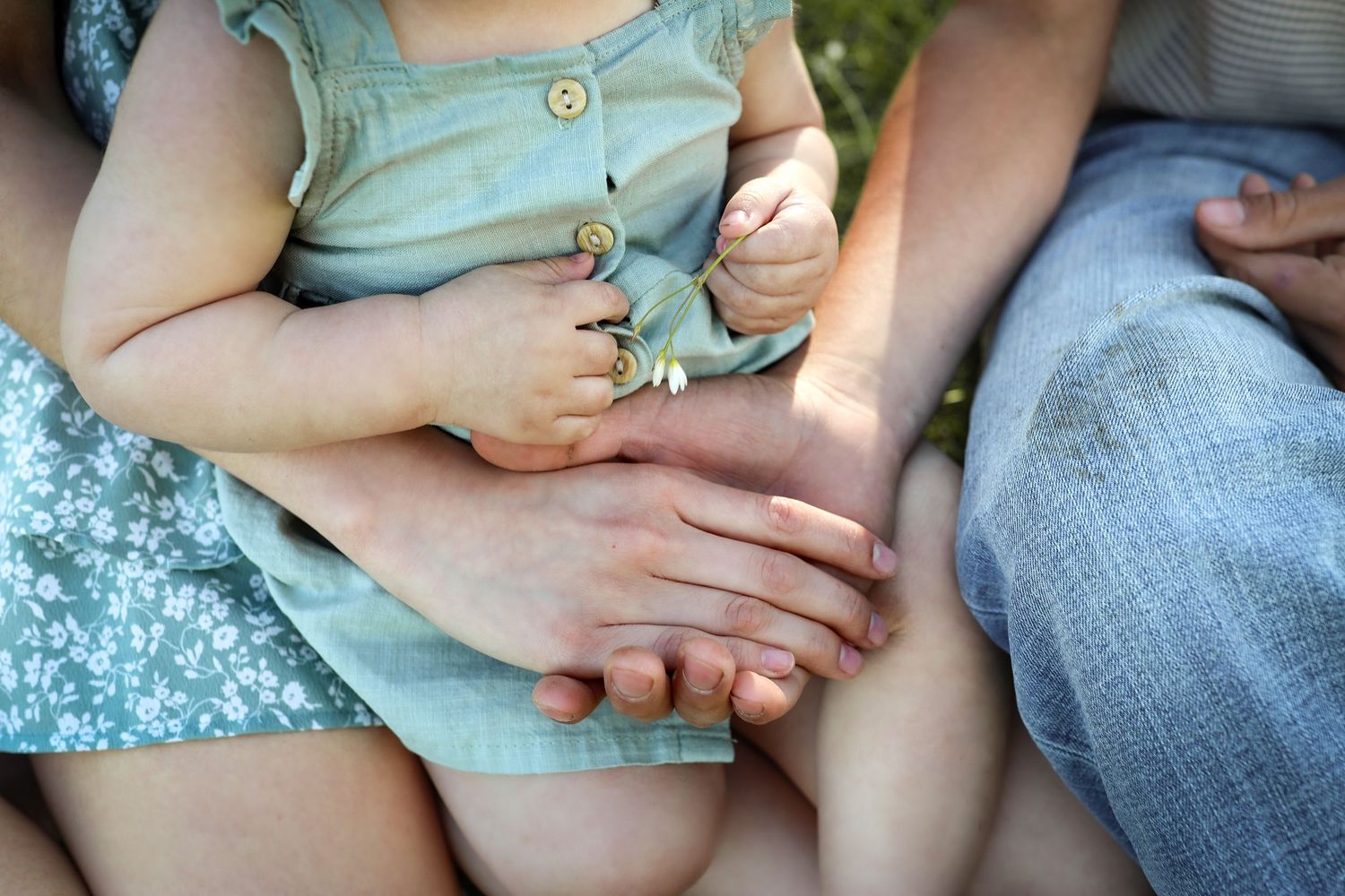 Close-up of adult hands gently holding a baby dressed in sage green romper while seated outdoors.
