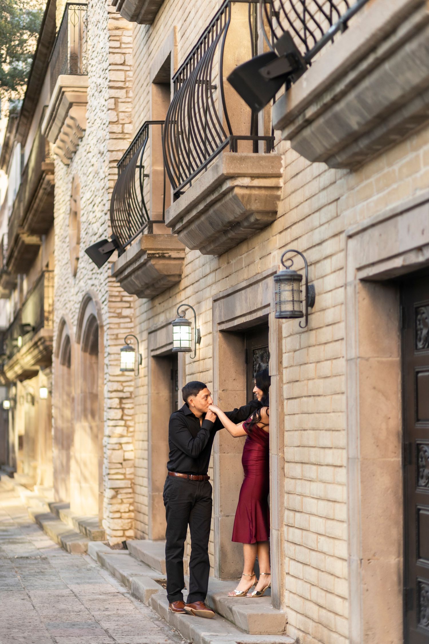 Man kissing woman's hand outside a stone building with balconies.