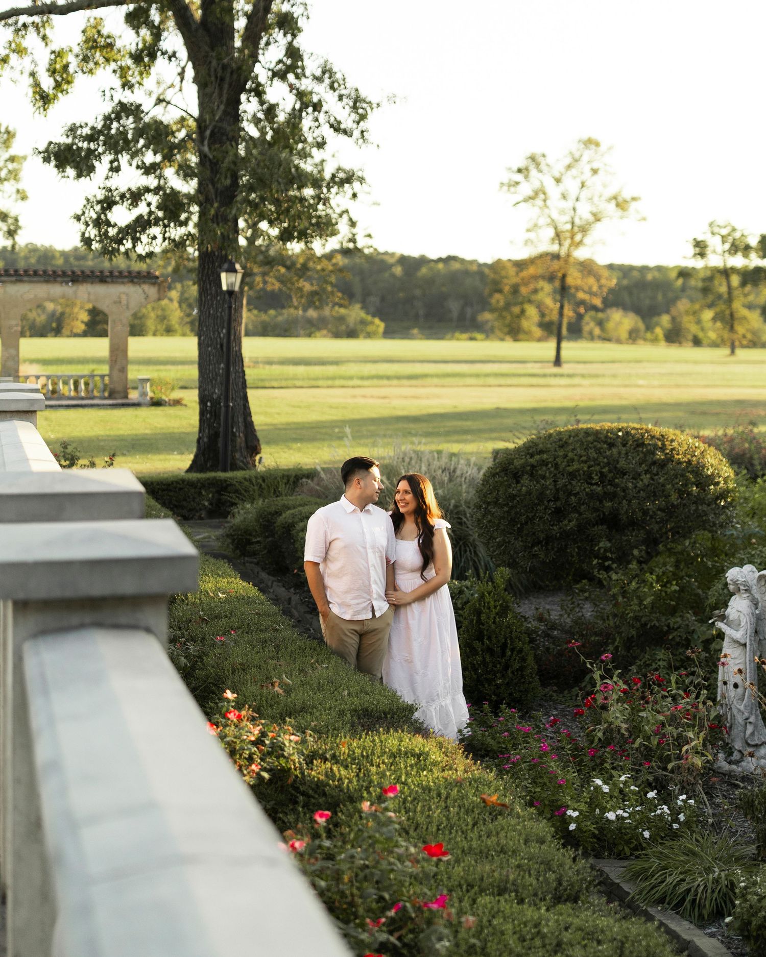 Couple standing in lush garden at sunset near stone balustrade.