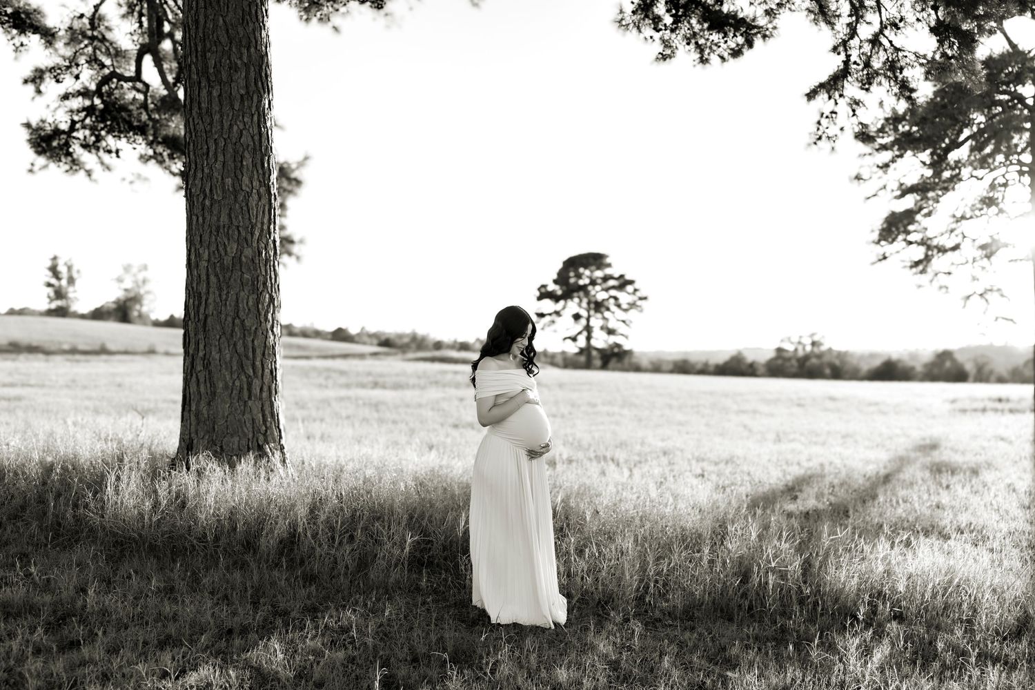 Pregnant woman in white dress standing under large tree in field, elegant black and white maternity photo.