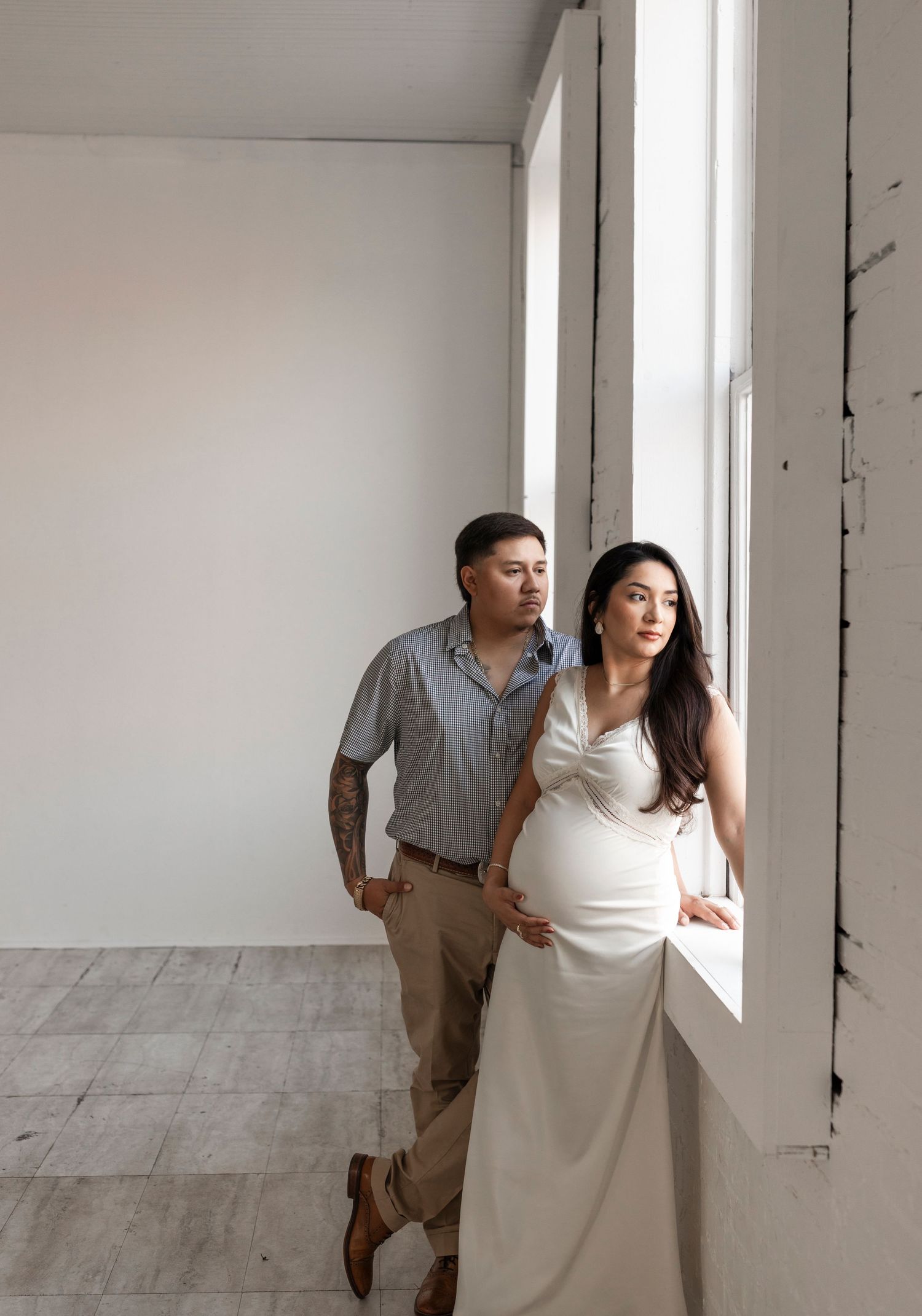 Couple standing by a large window in a white studio, woman in white gown holding belly.