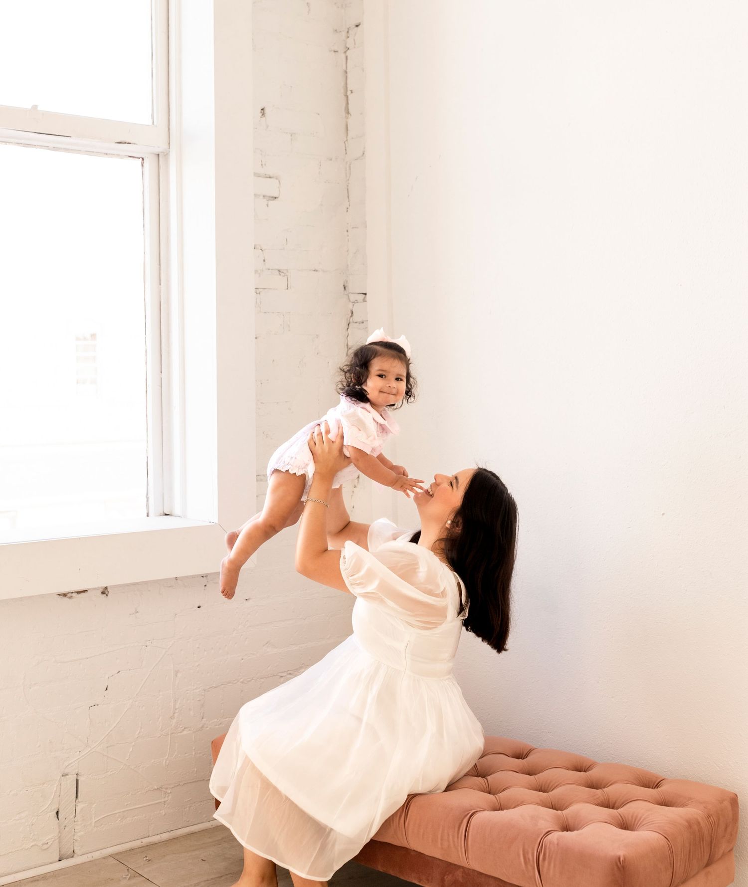 Mother in white dress lifting smiling toddler in bright white studio.