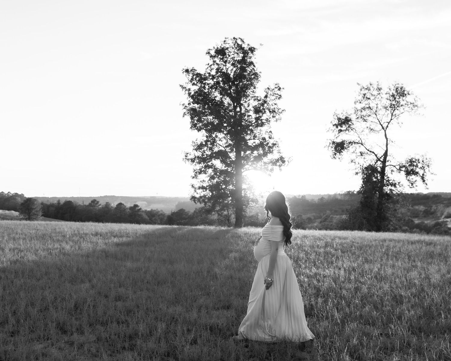 Black and white maternity photo of pregnant woman in white gown standing in field with sunset behind trees.