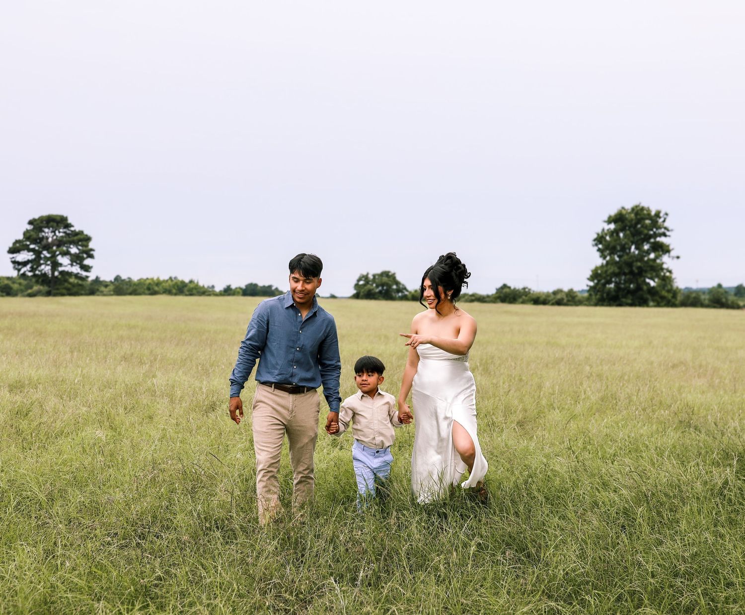 Family of three walking hand-in-hand through a lush green meadow, parents in casual attire with young child between them.