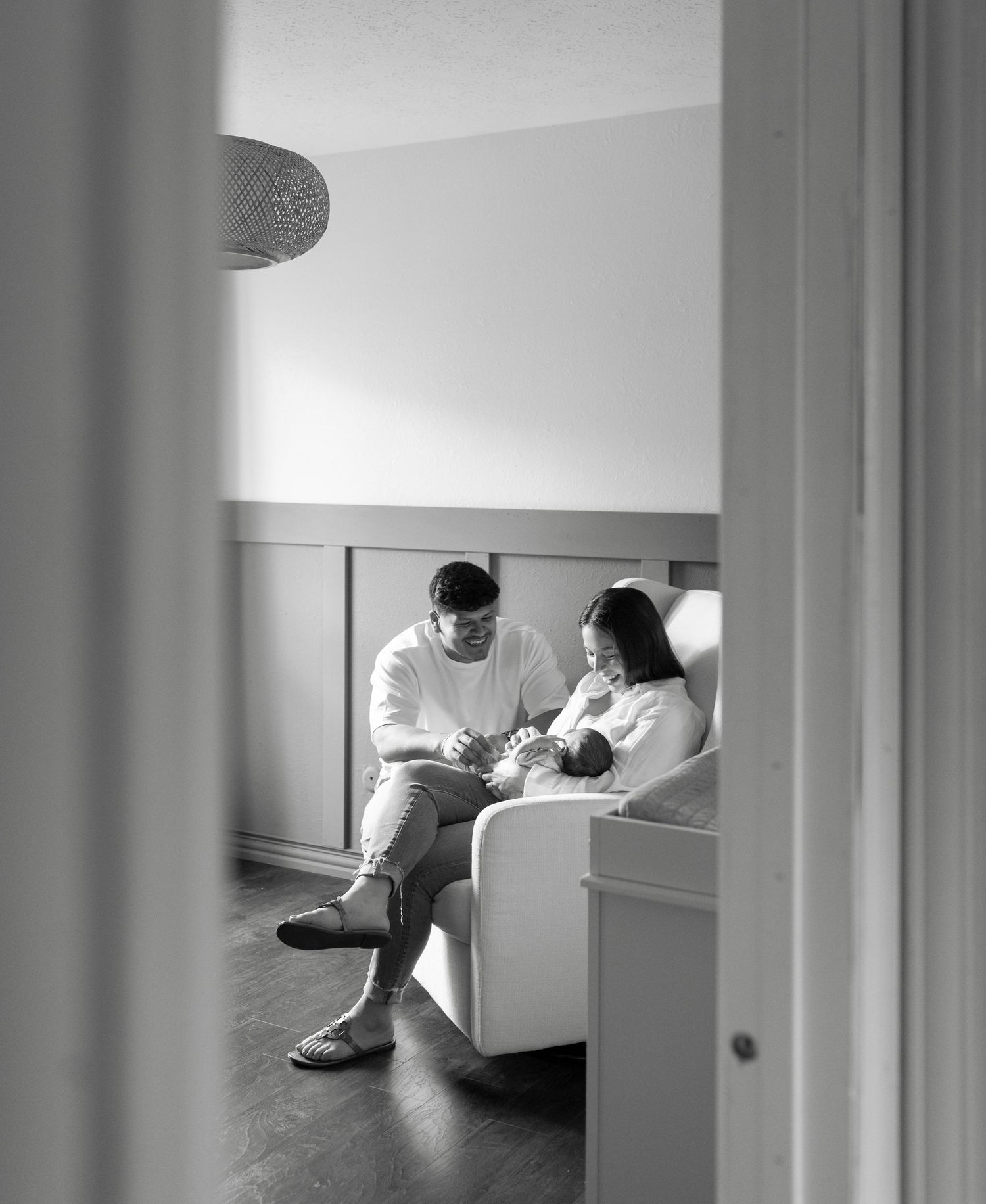 Black and white photo of new parents sitting together in a nursery chair with their newborn.